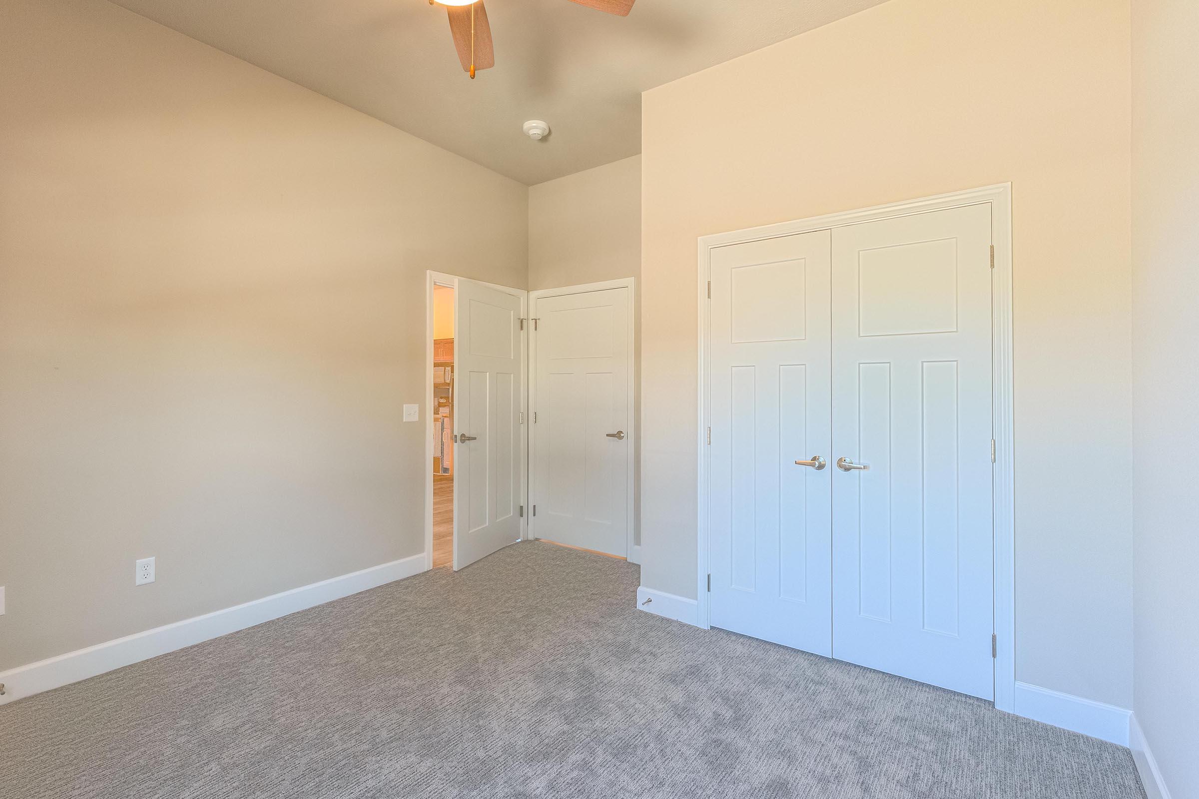 A bright and spacious room featuring neutral-colored walls and a plush carpet. Two closed white doors are visible, one leading to a closet and the other to another room. A ceiling fan is mounted above, and soft natural light filters in through a nearby doorway. The overall ambiance is clean and inviting.