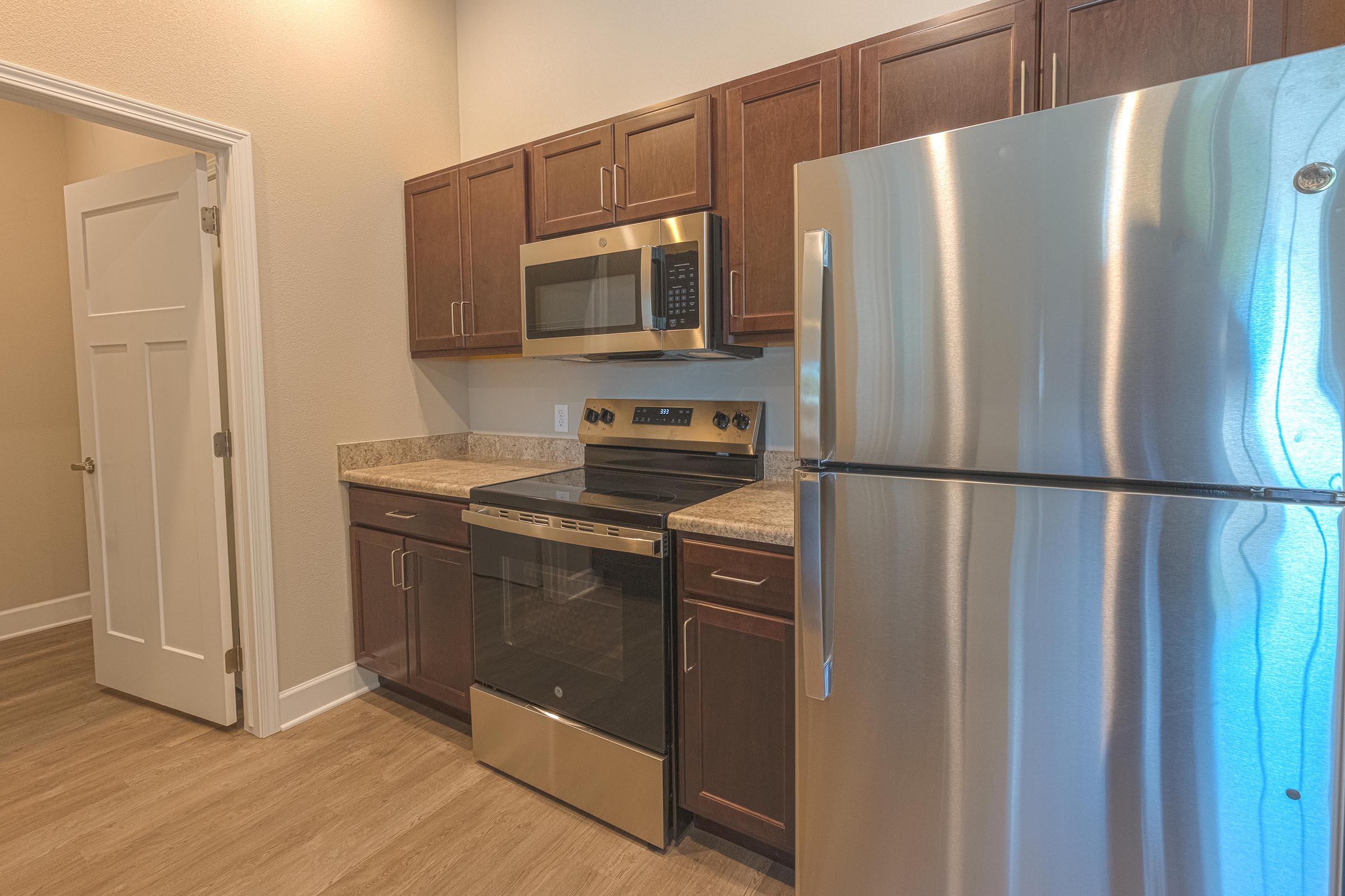 Modern kitchen featuring dark wood cabinets, granite countertops, stainless steel refrigerator, oven, and microwave. Bright and spacious layout with light-colored walls and a doorway leading to another room. The flooring is a light wood laminate.