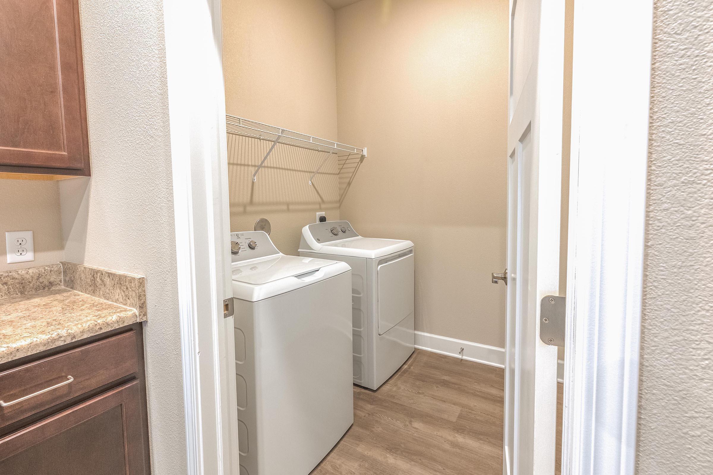 A laundry room featuring two white washing machines, a shelf above for storage, and light-colored walls. The flooring is a warm-toned laminate, and there are cabinets on the left side. The space is compact and well-organized, with a door partially open to the room.