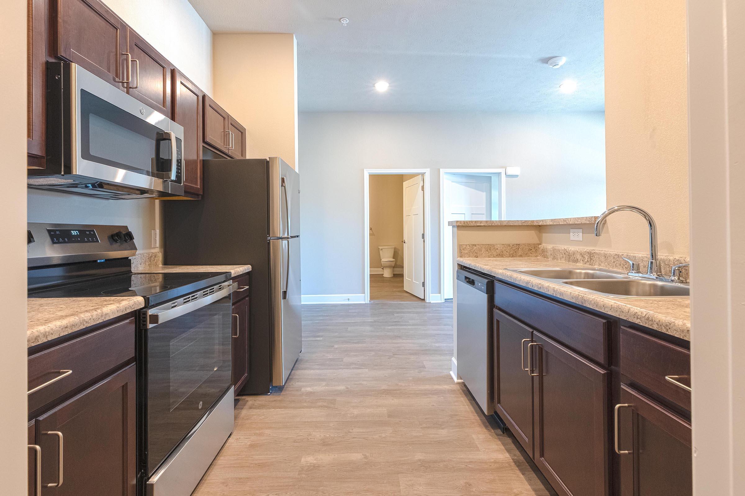 A modern kitchen featuring dark wooden cabinetry, stainless steel appliances including a microwave, oven, and refrigerator, and a double sink. The view leads to a hallway with a door opening to a bathroom. Bright and spacious with natural light filtering in.