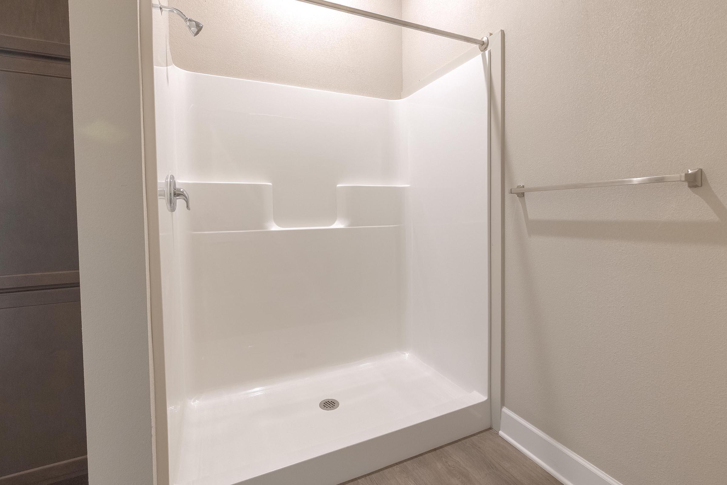 A clean and spacious shower stall with a white fiberglass interior. There is a showerhead installed on the left side, and a stainless steel towel bar mounted on the wall outside the enclosure. The flooring appears to be a light-colored laminate. The walls are neutral beige.