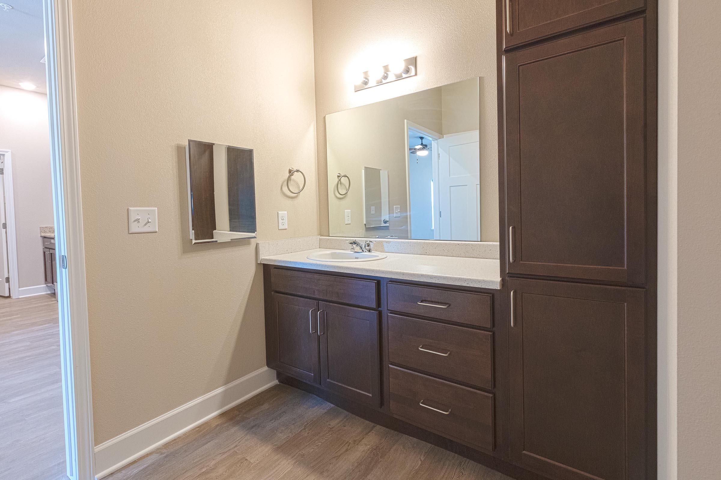 A modern bathroom featuring a light-colored wall, a large mirror above a dark wooden vanity with several drawers and a sink. There is an additional small mirror mounted on the wall. The floor has a light wood finish, and an open doorway leads to another room in the background.