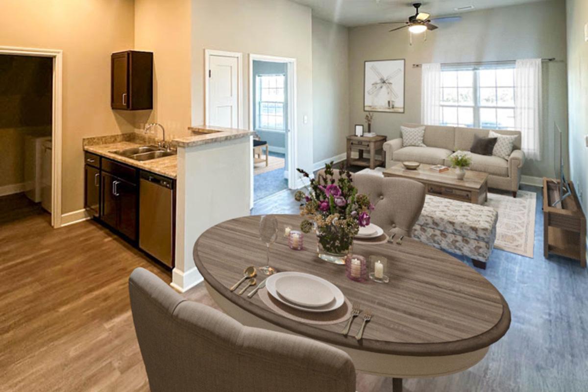 A modern, open living space featuring a cozy living room with a beige sofa, coffee table, and decorative plants, alongside a dining area with a round table set for dinner. The kitchen is visible with dark cabinets and granite countertops, while light walls and a large window provide a bright atmosphere.