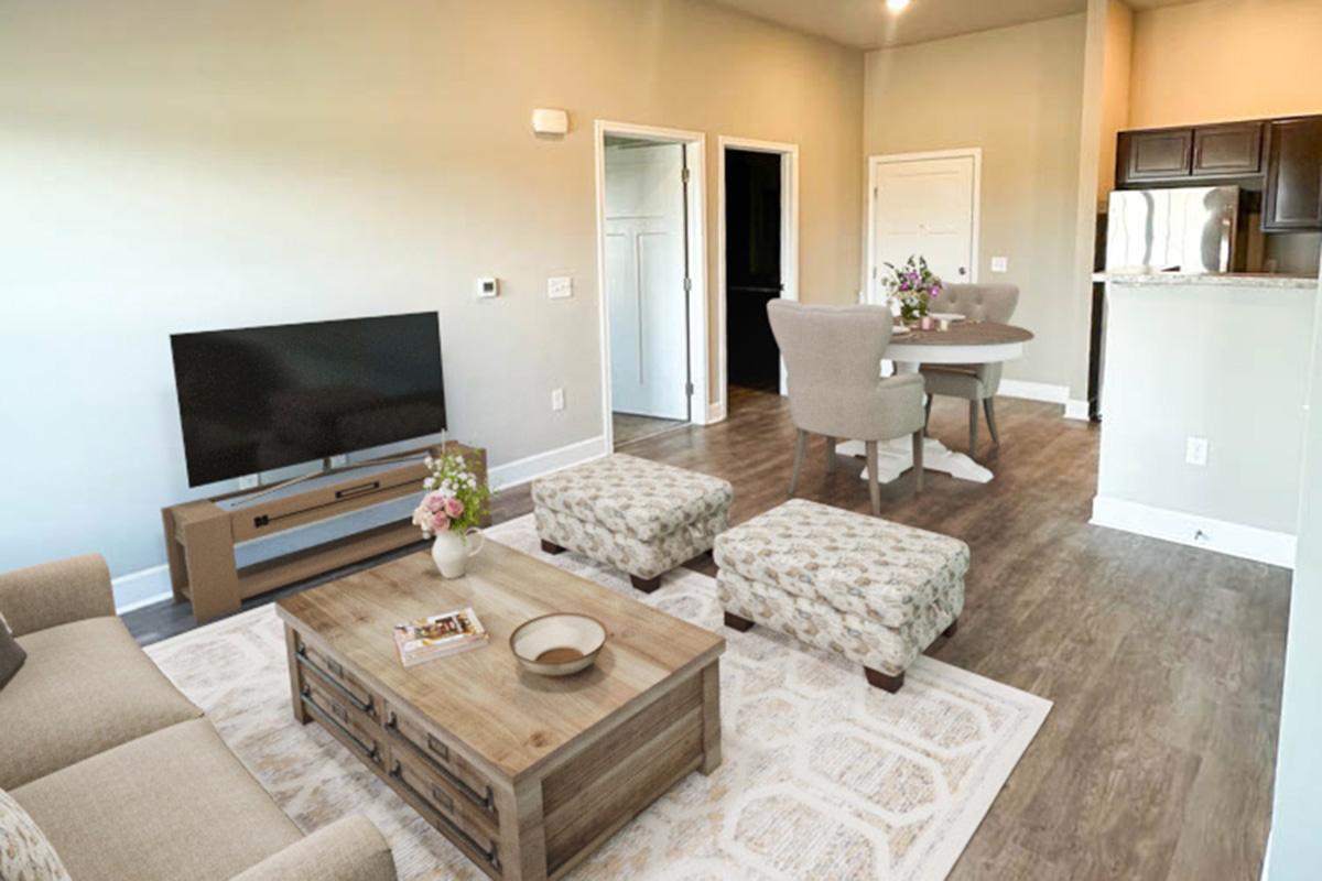 A modern living room featuring a light-colored sofa and a wooden coffee table with decorative items. Two patterned ottomans are in front of the sofa. A dining area with a round table and chairs is visible, along with a small kitchenette in the background. Soft lighting and neutral walls create a cozy atmosphere.