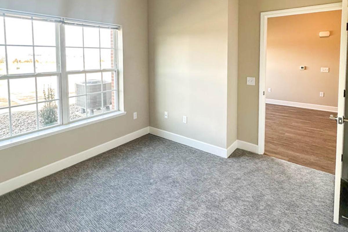 A spacious, empty room featuring a large window with a view, light-colored walls, and carpeted flooring. A doorway leads to another room in the background. The overall design gives a modern and fresh feel, suitable for various uses.