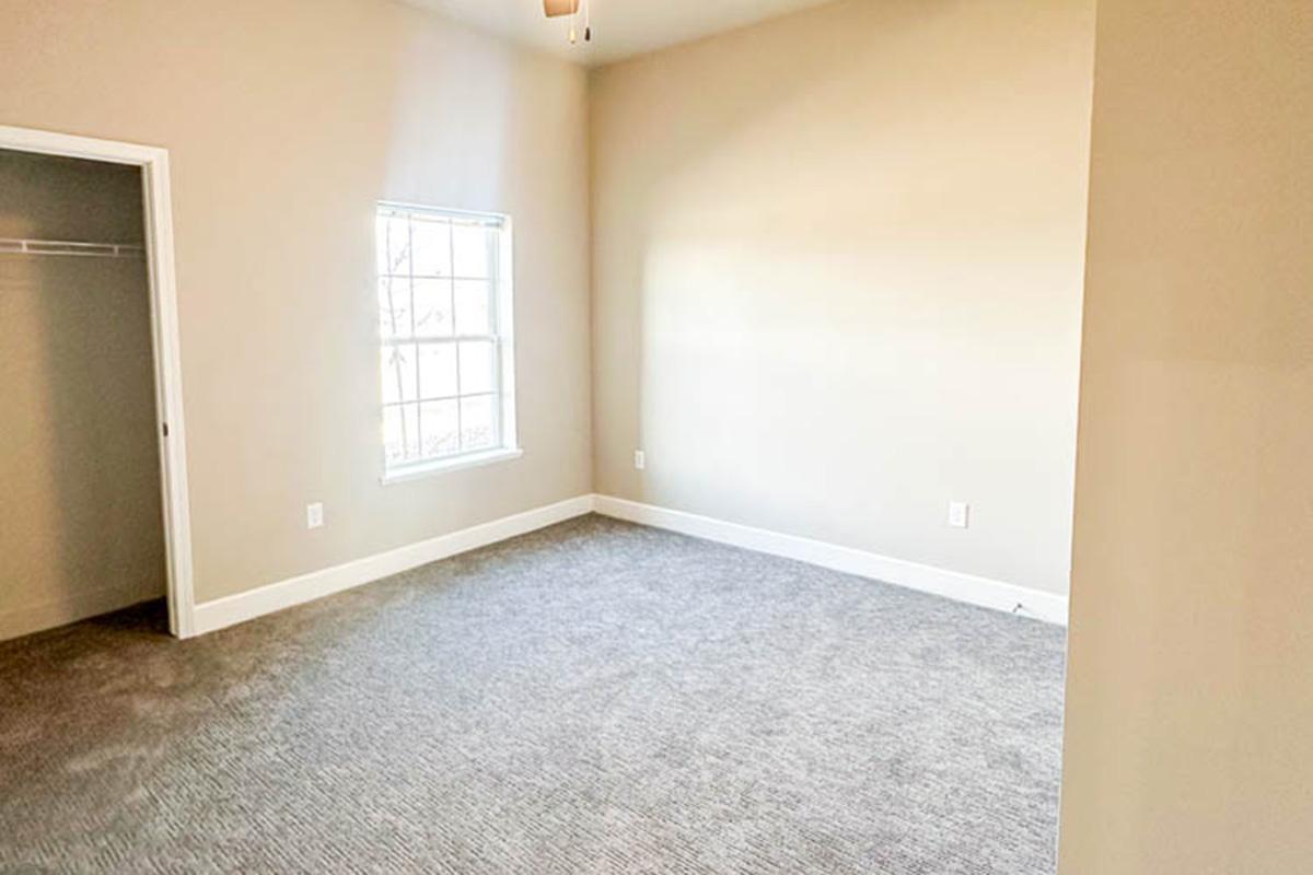 Empty room with light beige walls and gray carpet. There is a closet to the left and a window on the right side allowing natural light to enter. The space appears bright and airy, with no furniture present.