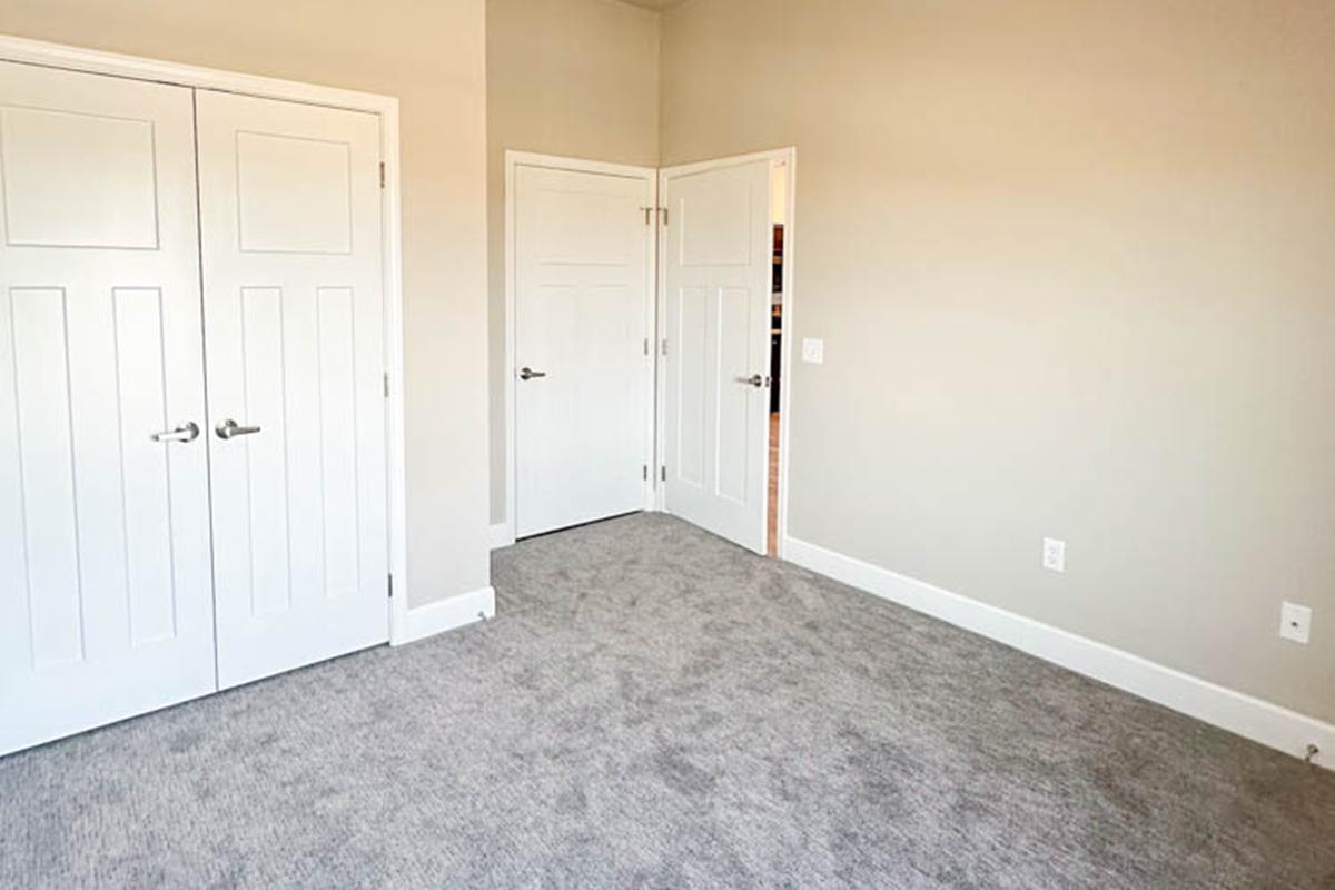 A clean, empty room featuring light gray walls and a plush carpet. Two double doors on the left lead to a closet, and another door on the right opens to the hallway. Natural light gently illuminates the space, creating a bright and airy atmosphere.