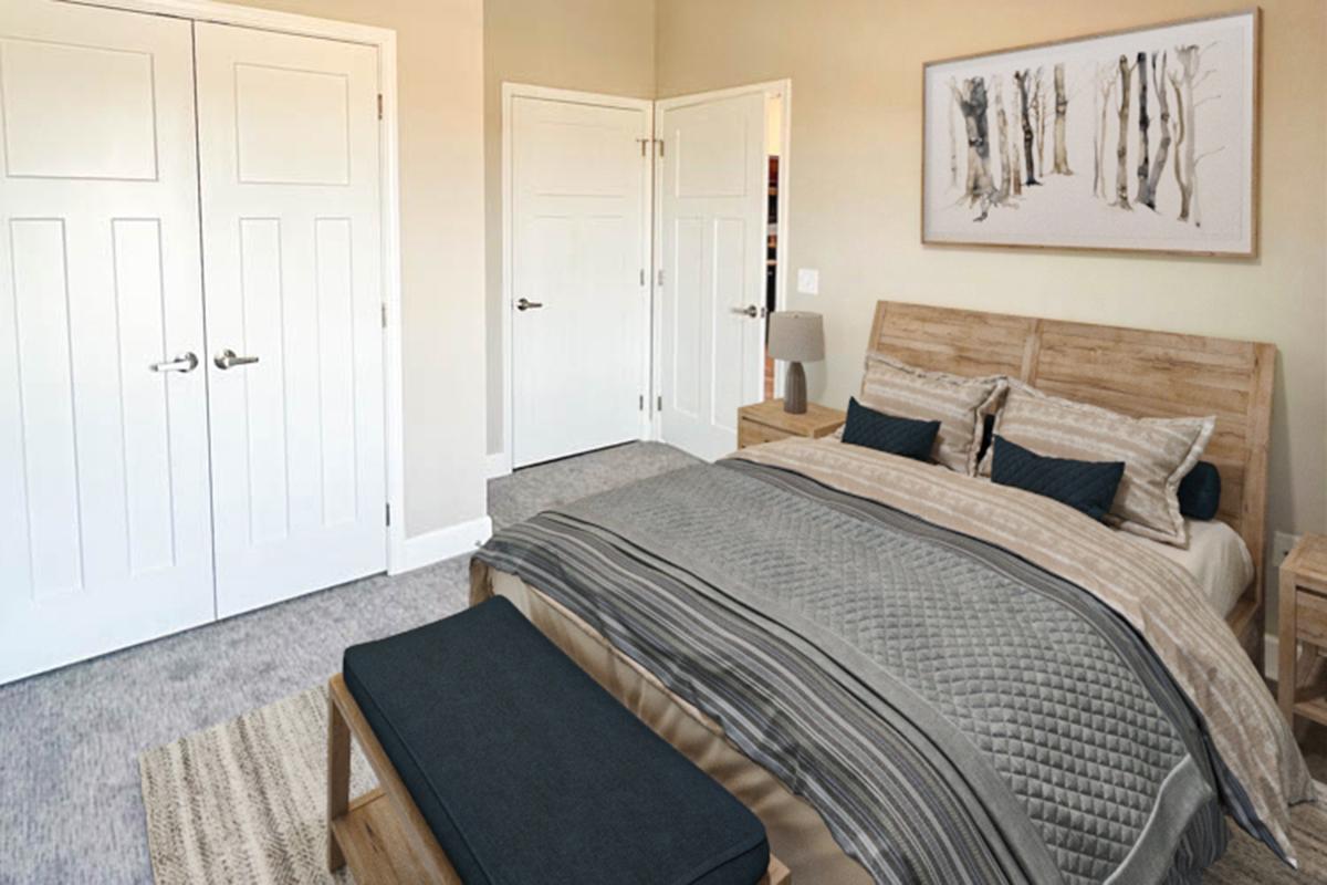 A cozy bedroom featuring a wooden bed frame with layered bedding in neutral tones. There's a bench at the foot of the bed and two nightstands with decorative lamps. The walls are painted a soft color, and artwork of trees hangs above the bed. Two white closet doors are visible, adding to the room's inviting atmosphere.