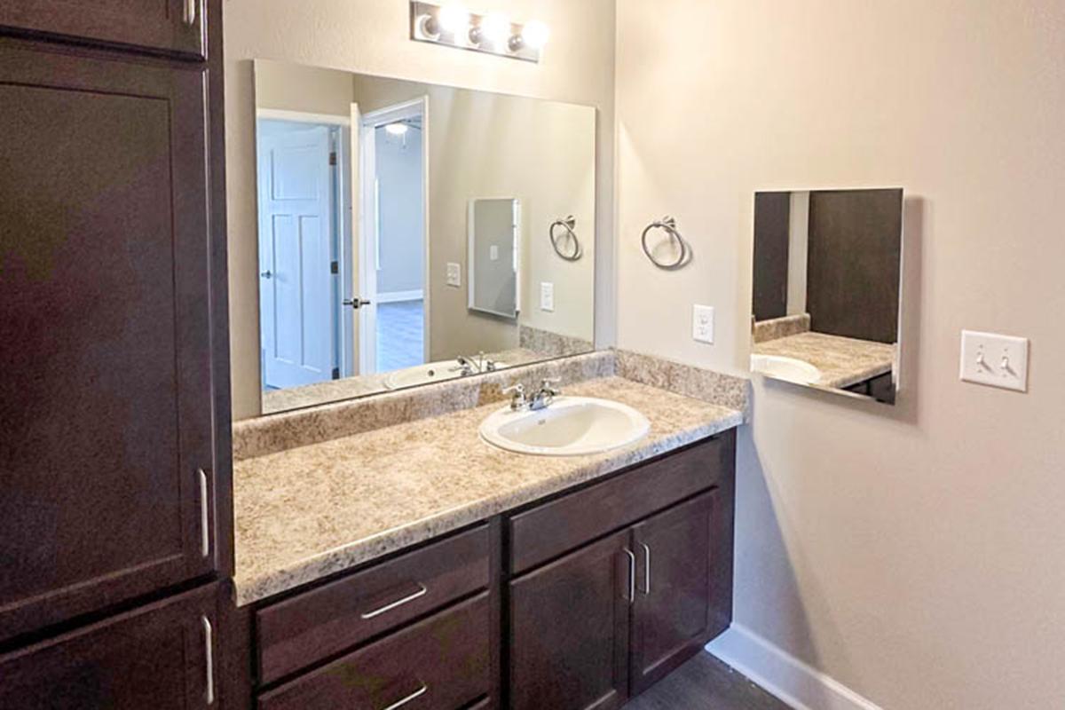 A modern bathroom with a double vanity featuring a beige countertop and dark wood cabinetry. Mirrors are mounted above the sinks, and the space is well-lit by an overhead light fixture. A door in the background leads to another room. The walls are painted a soft color, enhancing the bright, clean aesthetic.