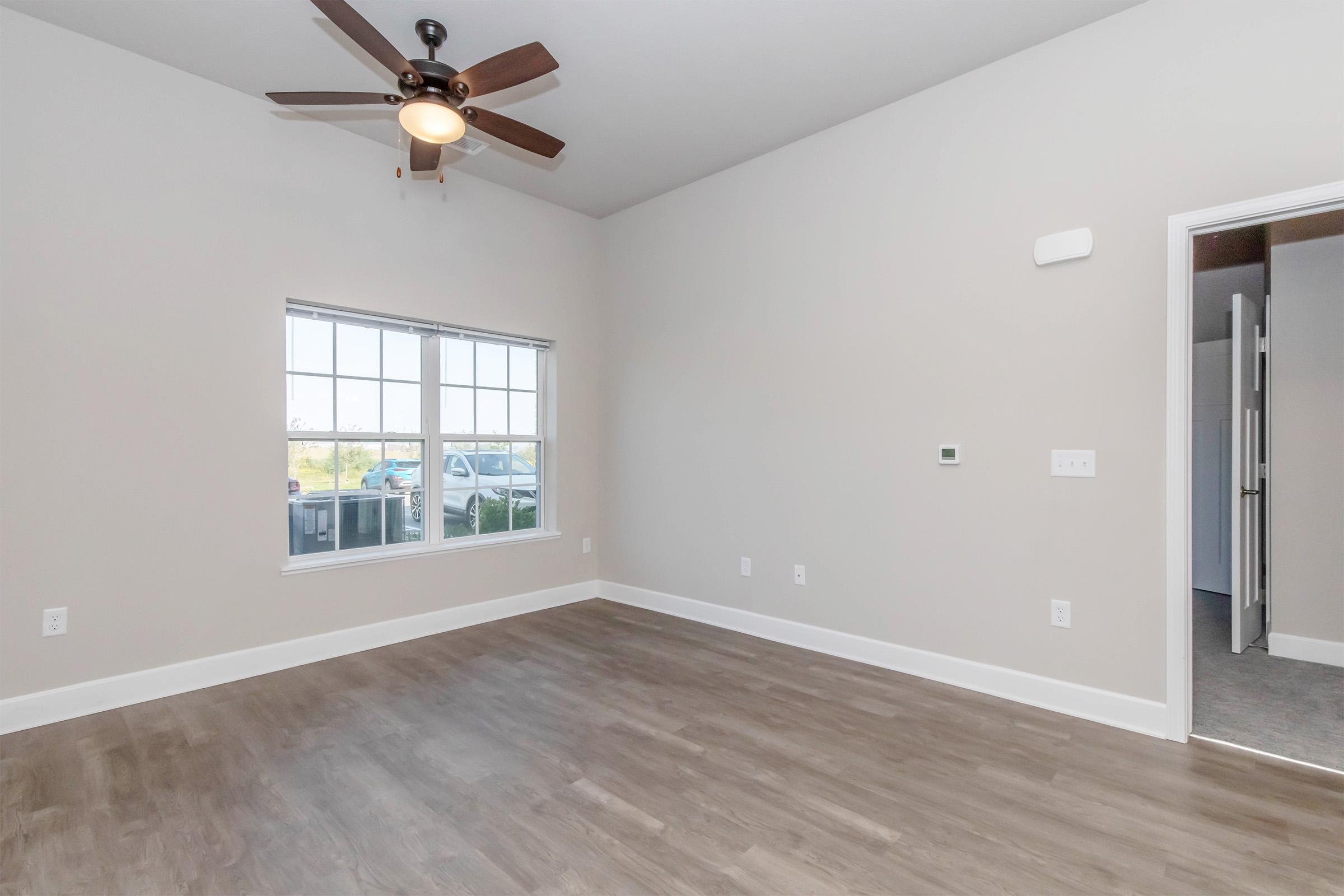 A well-lit, spacious room featuring light-colored walls and brown wood-like flooring. A ceiling fan is visible above, and a large window allows natural light to enter the space. There is a thermostat on the wall, and a doorway leads to another room. The overall atmosphere is clean and modern.