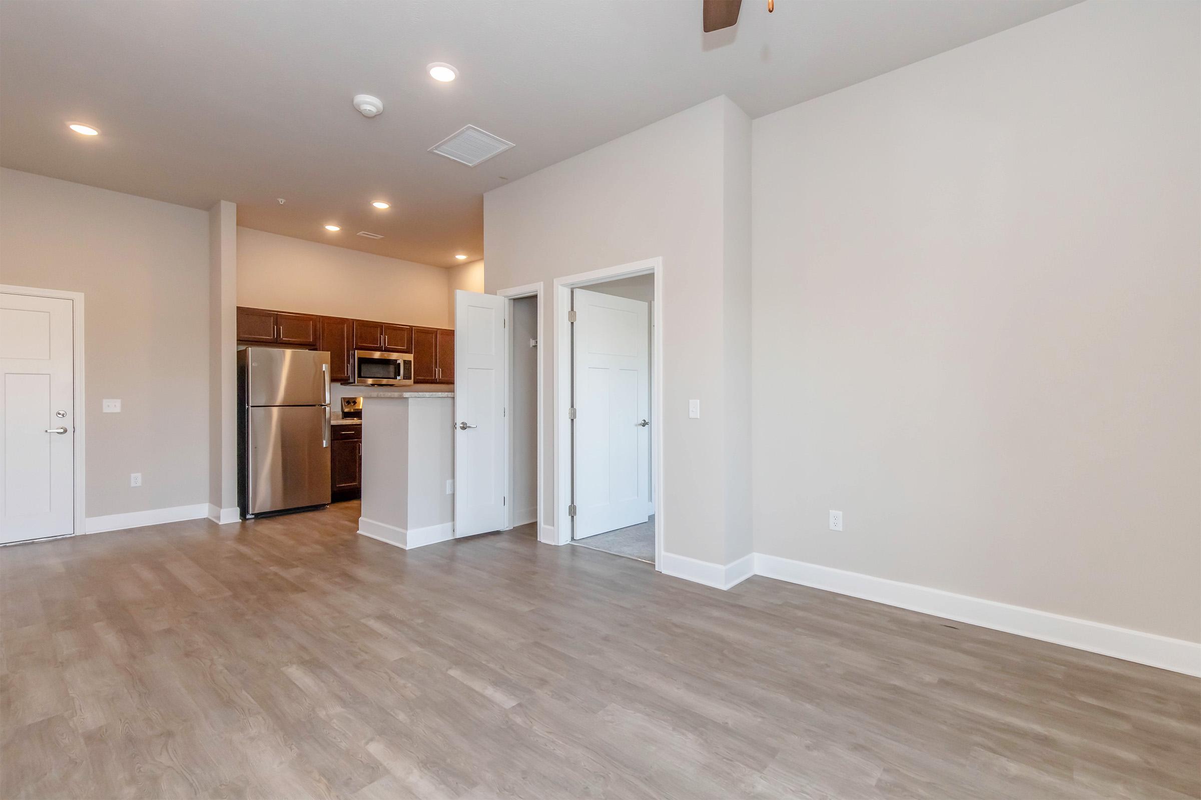 Spacious modern living area with light-colored walls and wood-style flooring. The kitchen features stainless steel appliances and a small bar area. Visible doors lead to a bathroom and bedrooms, with ample natural light coming through. Ceiling fan installed for airflow.