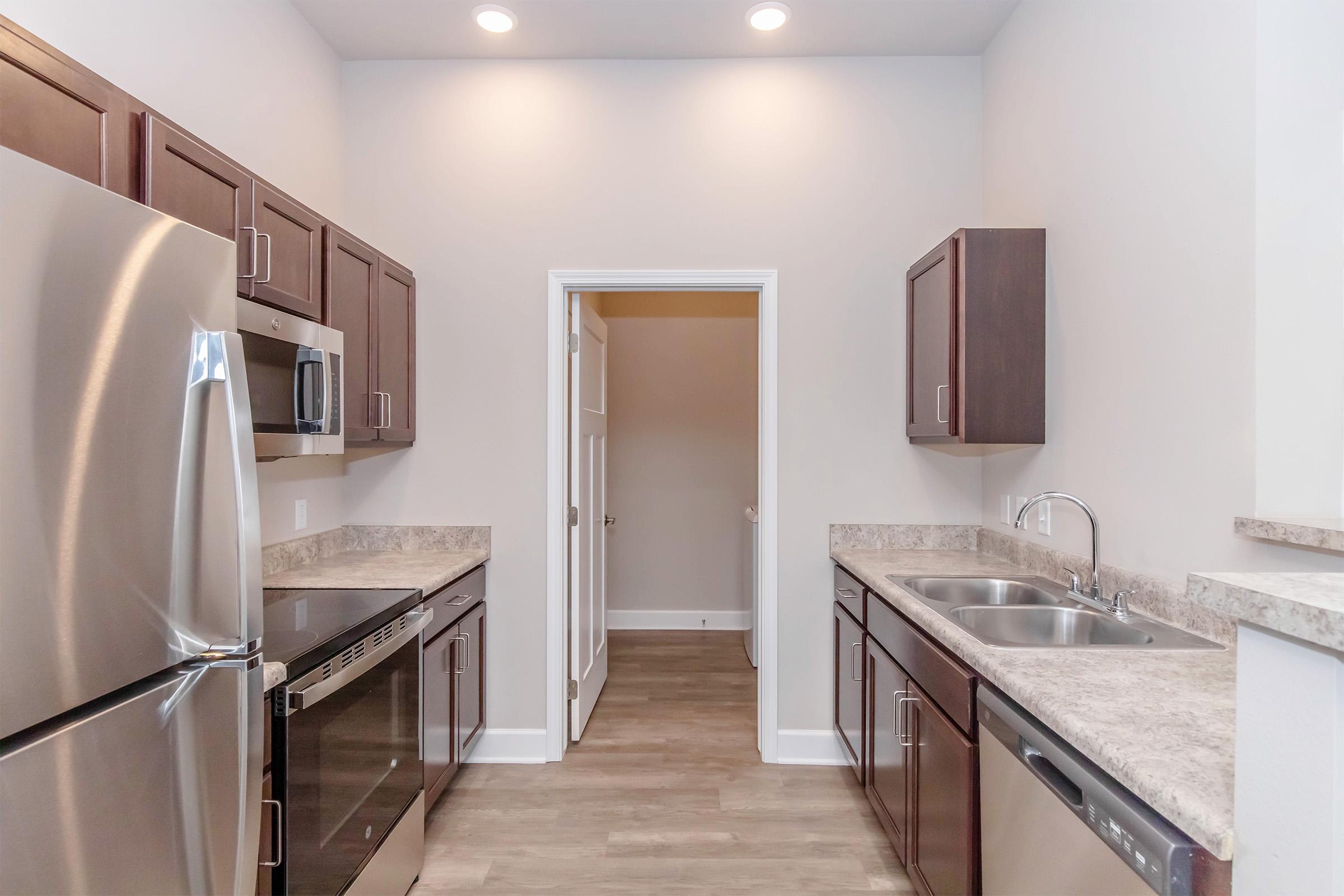 A modern kitchen featuring dark wooden cabinets, stainless steel appliances, and a granite countertop. The space is well-lit with recessed lighting and includes a double sink. A door leads to a pantry or closet area, with light-colored walls and a neutral-toned floor.