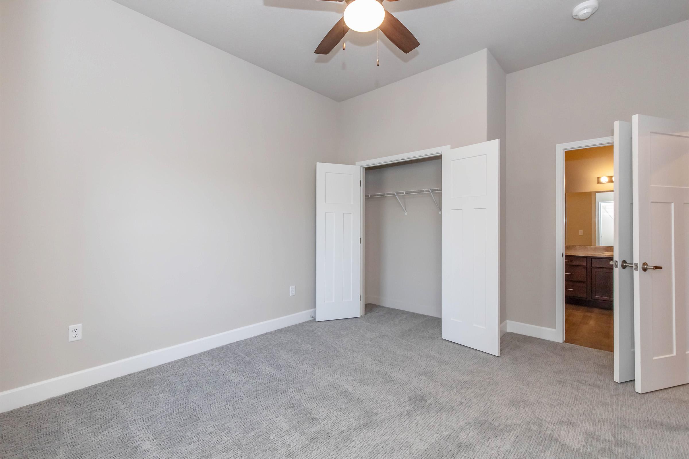 A spacious, empty bedroom with light gray carpeting and beige walls. Two white doors are open, revealing a closet with a hanging rail. A ceiling fan is visible above, and light enters from an adjoining room. The overall atmosphere is bright and inviting.