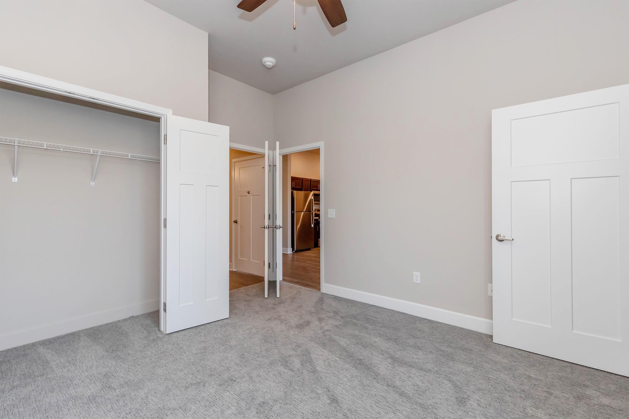 A spacious, light-colored room with carpet flooring, featuring open double doors leading to another room. One wall has a closet with shelving, and the ceiling has a ceiling fan. The room has neutral-colored walls, creating a bright and airy atmosphere.