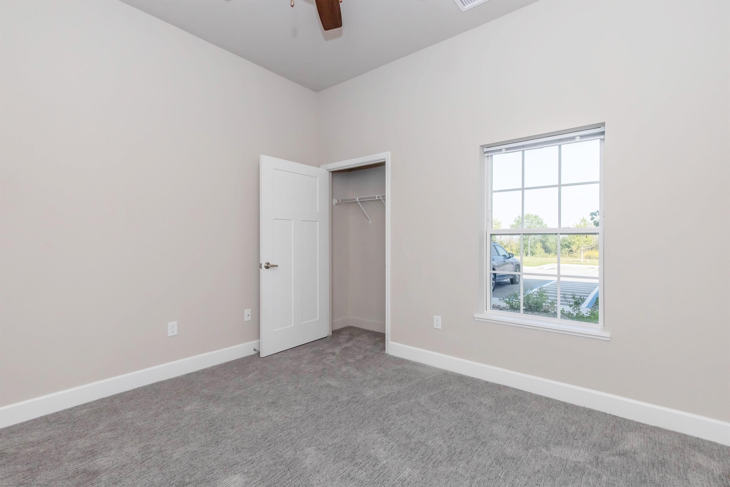 A vacant room with light gray carpet and neutral walls. There's a white door leading to a closet and a window with a view outside. The room appears bright and spacious, with natural light coming in through the window.