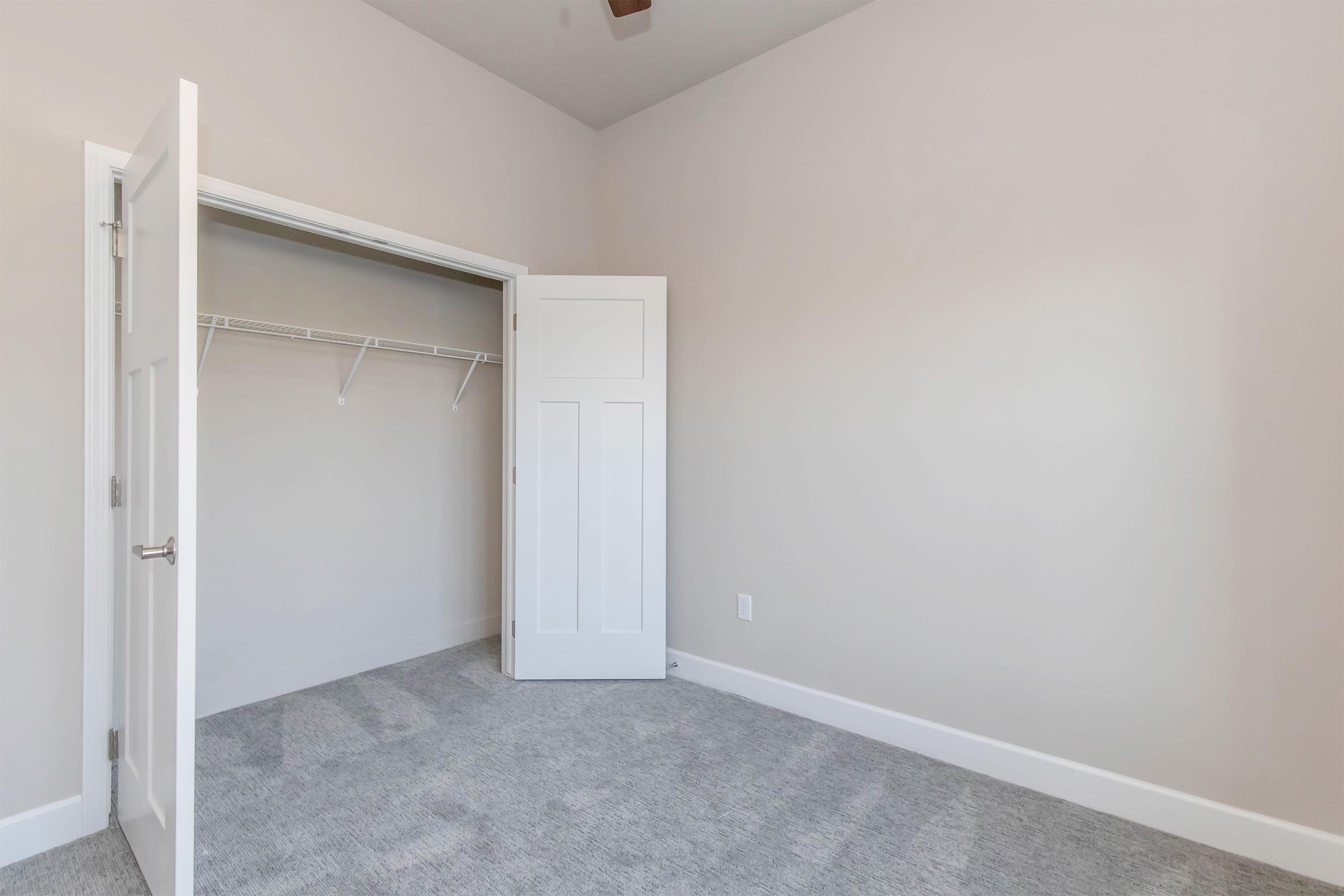 A vacant room with light-colored walls and carpeted flooring. A closet with a white sliding door is visible, featuring a simple design and a single light fixture on the ceiling. The space appears clean and well-lit, suggesting a minimalist aesthetic.