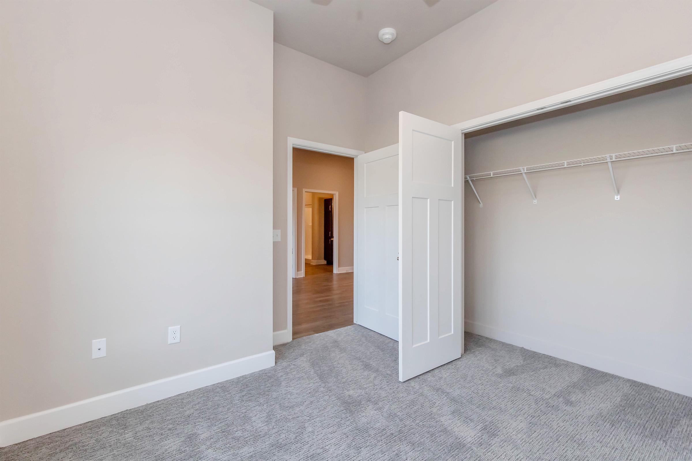 A spacious, light-colored room featuring an open closet with a white door. The walls are painted in a soft tone, and the floor is covered with light grey carpet. A doorway leads to another room in the background, indicating additional living space. The ceiling is fitted with a ceiling fan.