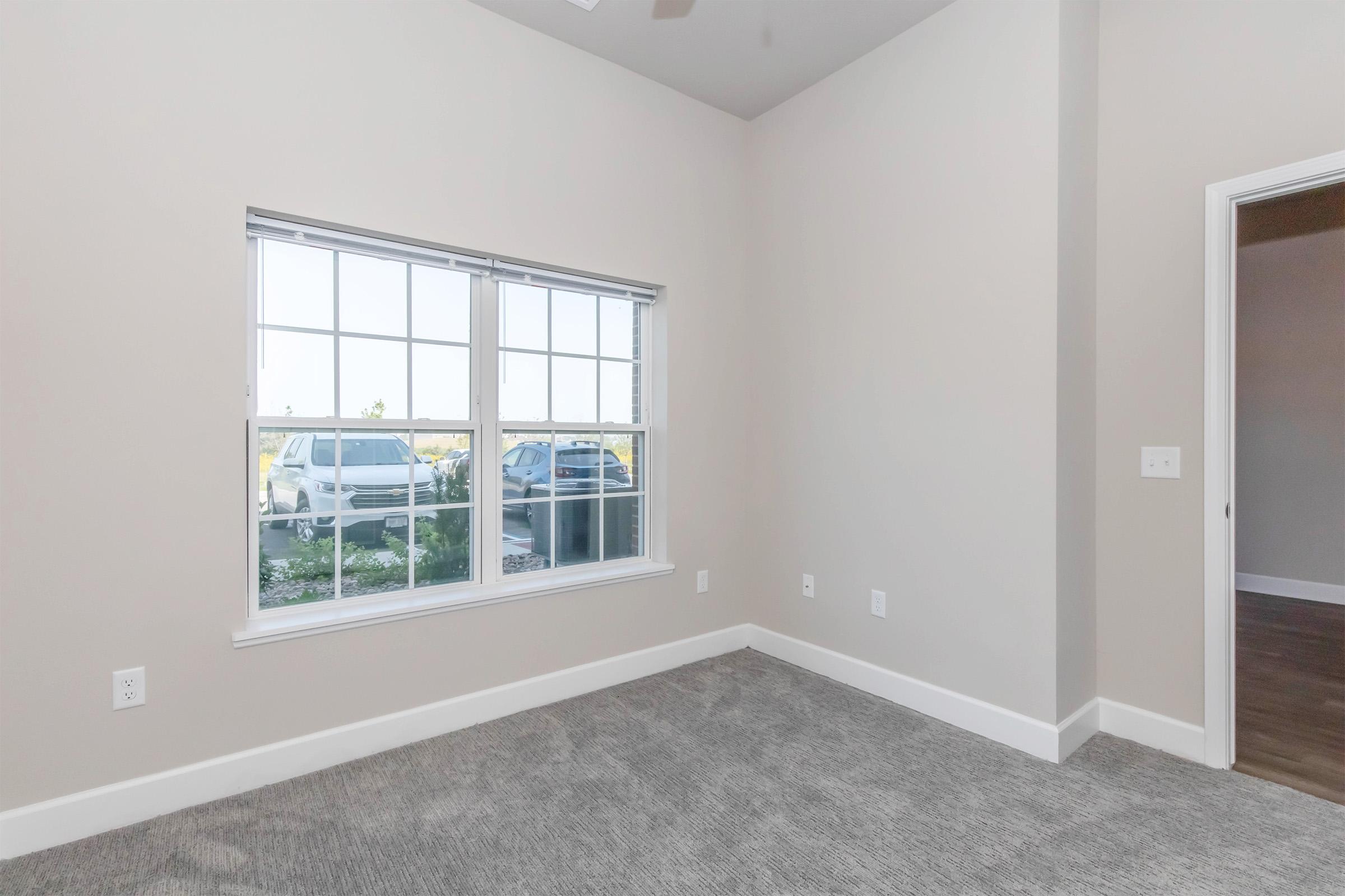 A vacant room with light gray walls and carpet flooring. It features a large window with white trim, allowing natural light to enter. Outside the window, parked cars are visible. The room has a ceiling fan and a doorway leading to another area, suggesting an open layout.