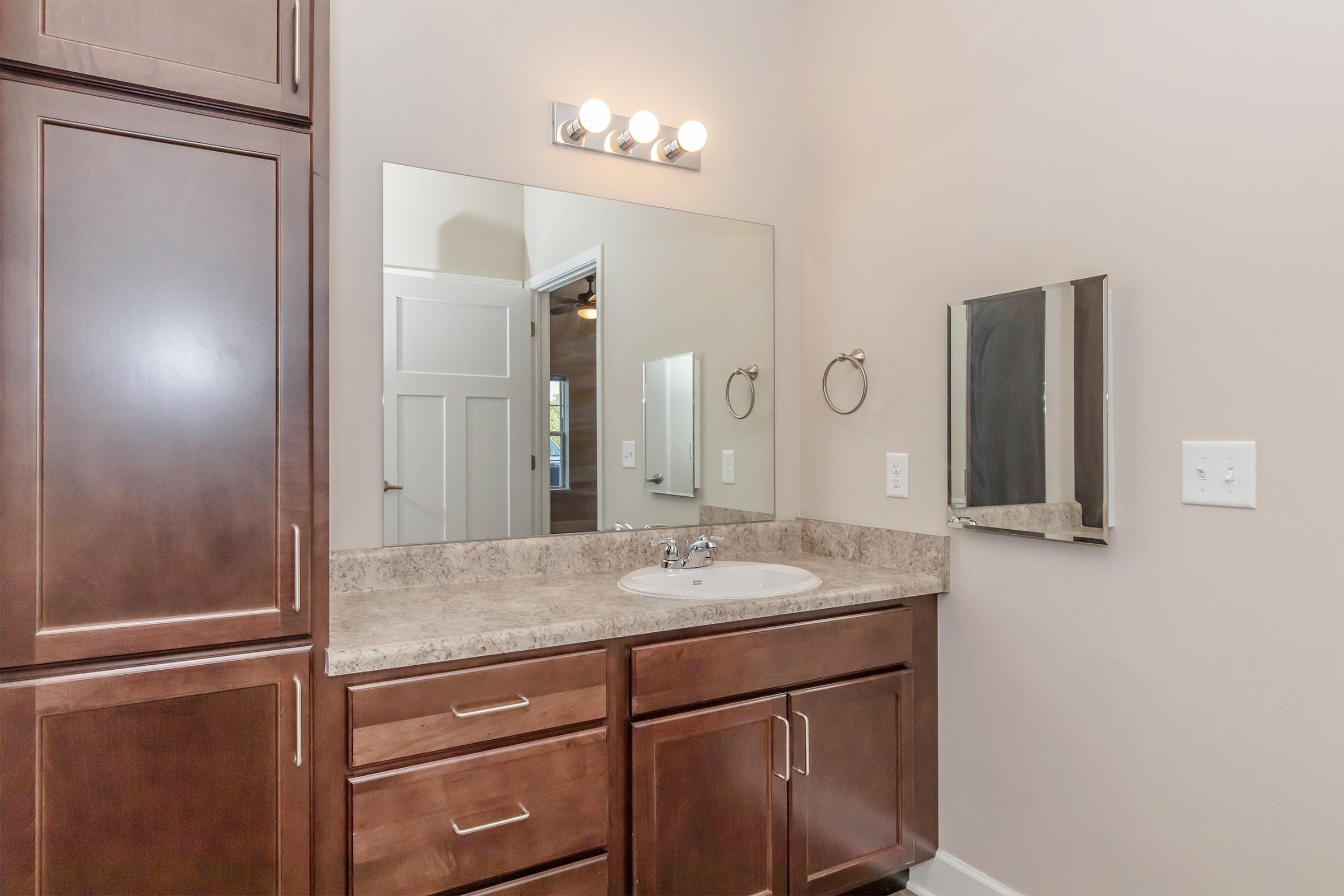 A modern bathroom featuring a brown wood vanity with a sink, a large mirror above, and stylish light fixtures. The walls are painted a neutral color, and there are towel holders next to the mirror. The door to another room is visible in the background, indicating a spacious layout.