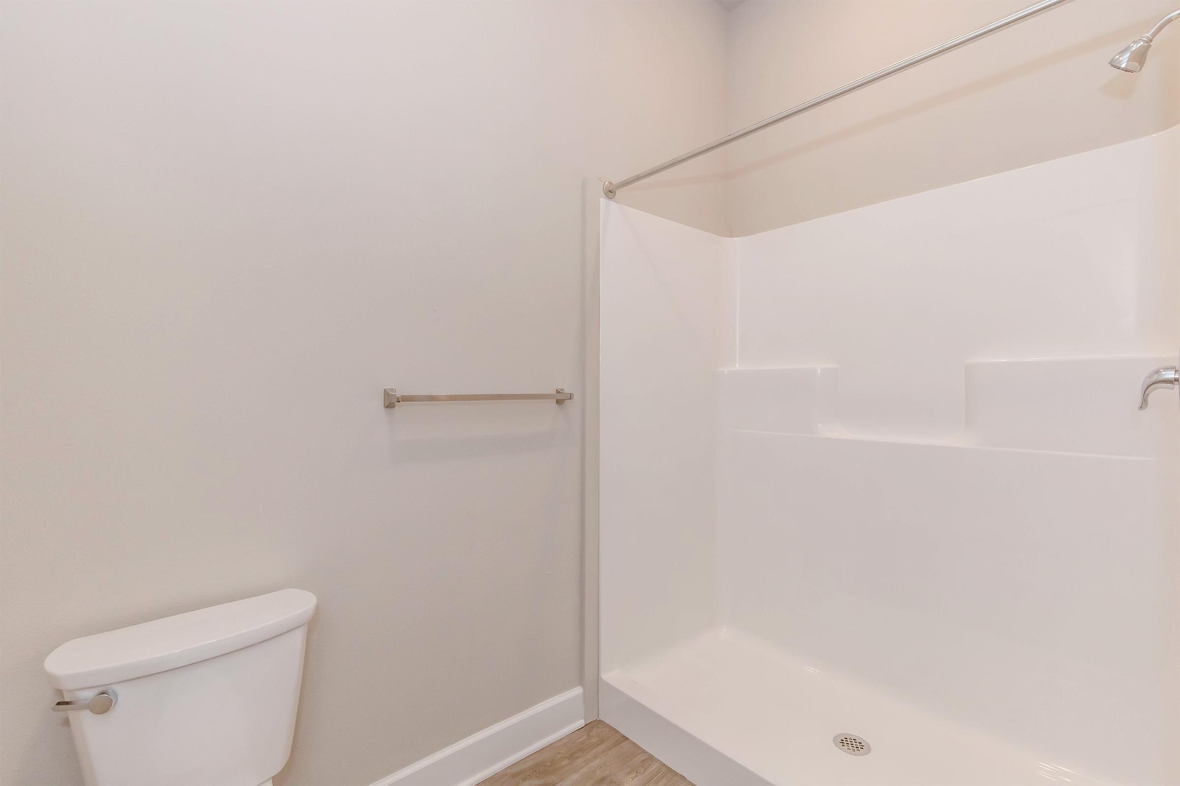A simple, clean bathroom featuring a white shower stall with a sleek glass door, a toilet beside it, and light-colored walls. The floor is finished with neutral-toned tiles, creating a modern and minimalistic look.