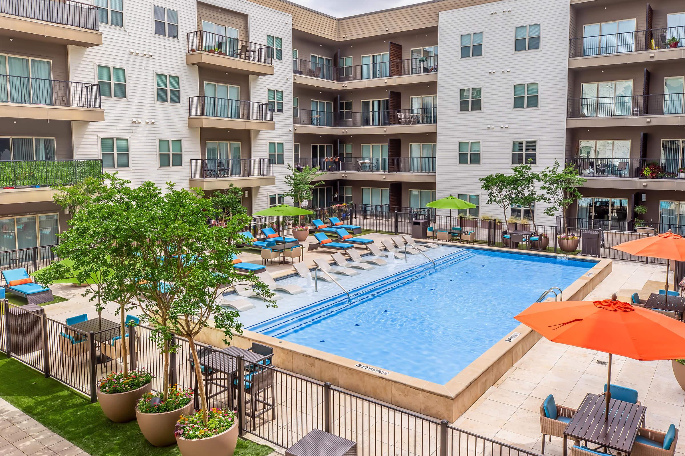 A spacious outdoor pool area surrounded by lounge chairs and green umbrellas, with modern apartment buildings in the background. The scene includes landscaped greenery, patio seating with tables, and vibrant orange umbrellas, creating a relaxing communal space. Bright and inviting atmosphere.