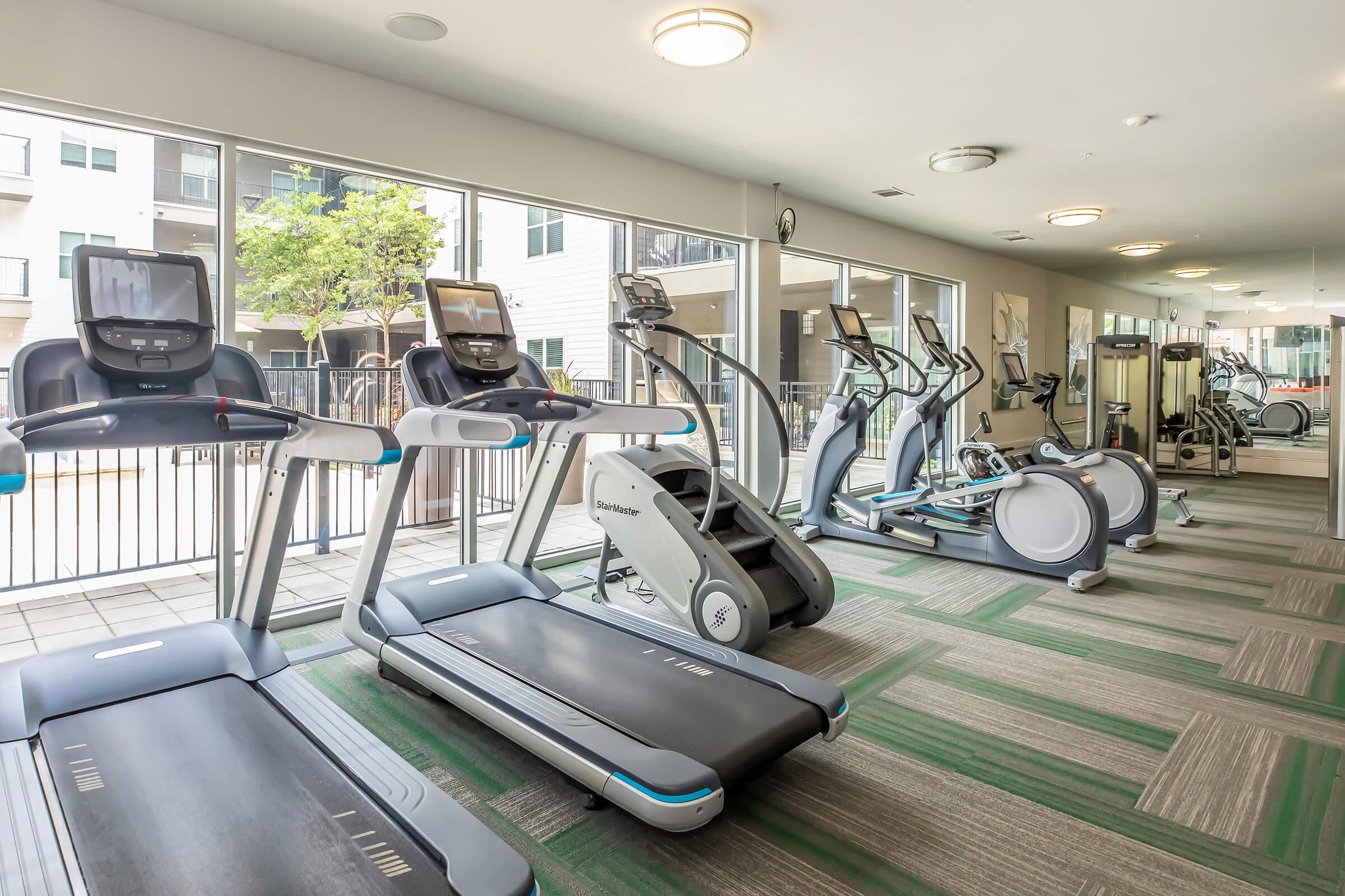 A modern fitness center featuring several exercise machines, including treadmills, ellipticals, and stationary bikes. The space has large windows allowing natural light in, with a view of outdoor greenery. The flooring is a green and gray patterned carpet, creating a clean, inviting workout environment.