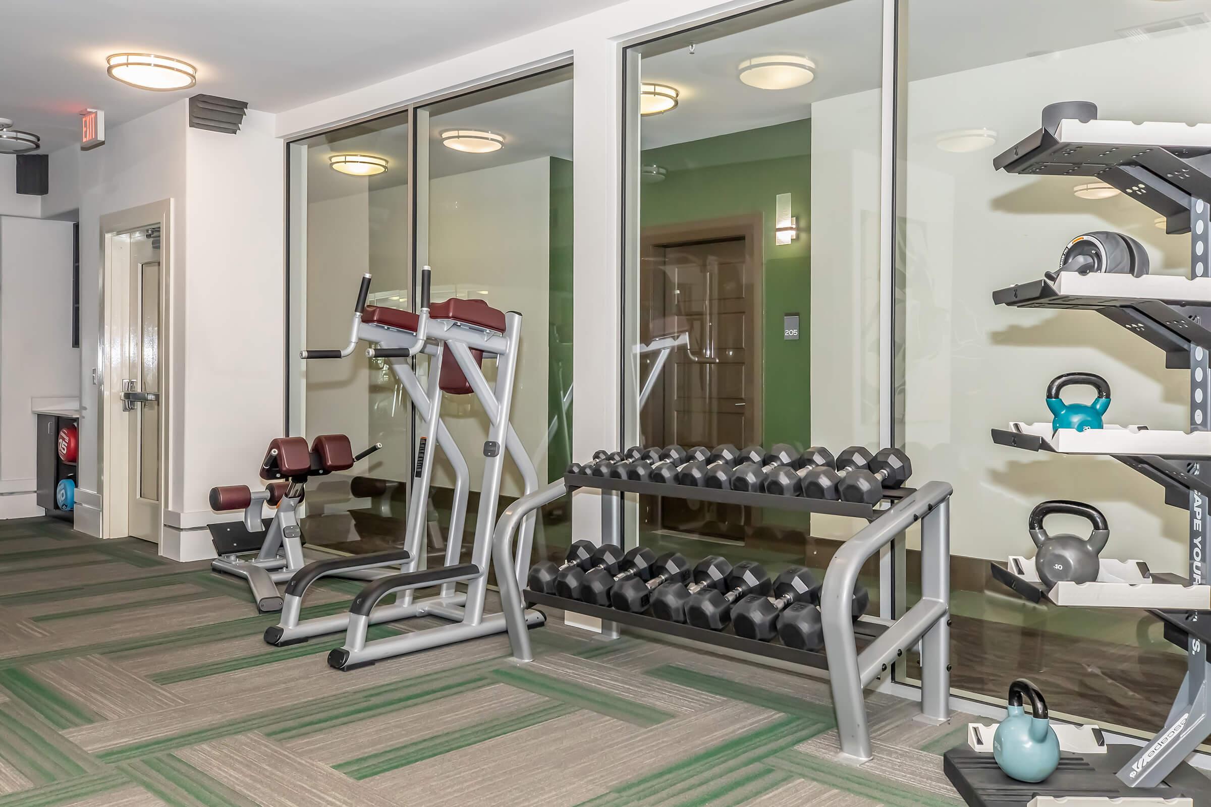 A modern gym interior featuring a variety of fitness equipment. There are weightlifting benches, dumbbells lined on racks, and exercise machines, all situated in a well-lit space with large windows, providing a view of the outside. The flooring is patterned with green and gray designs.