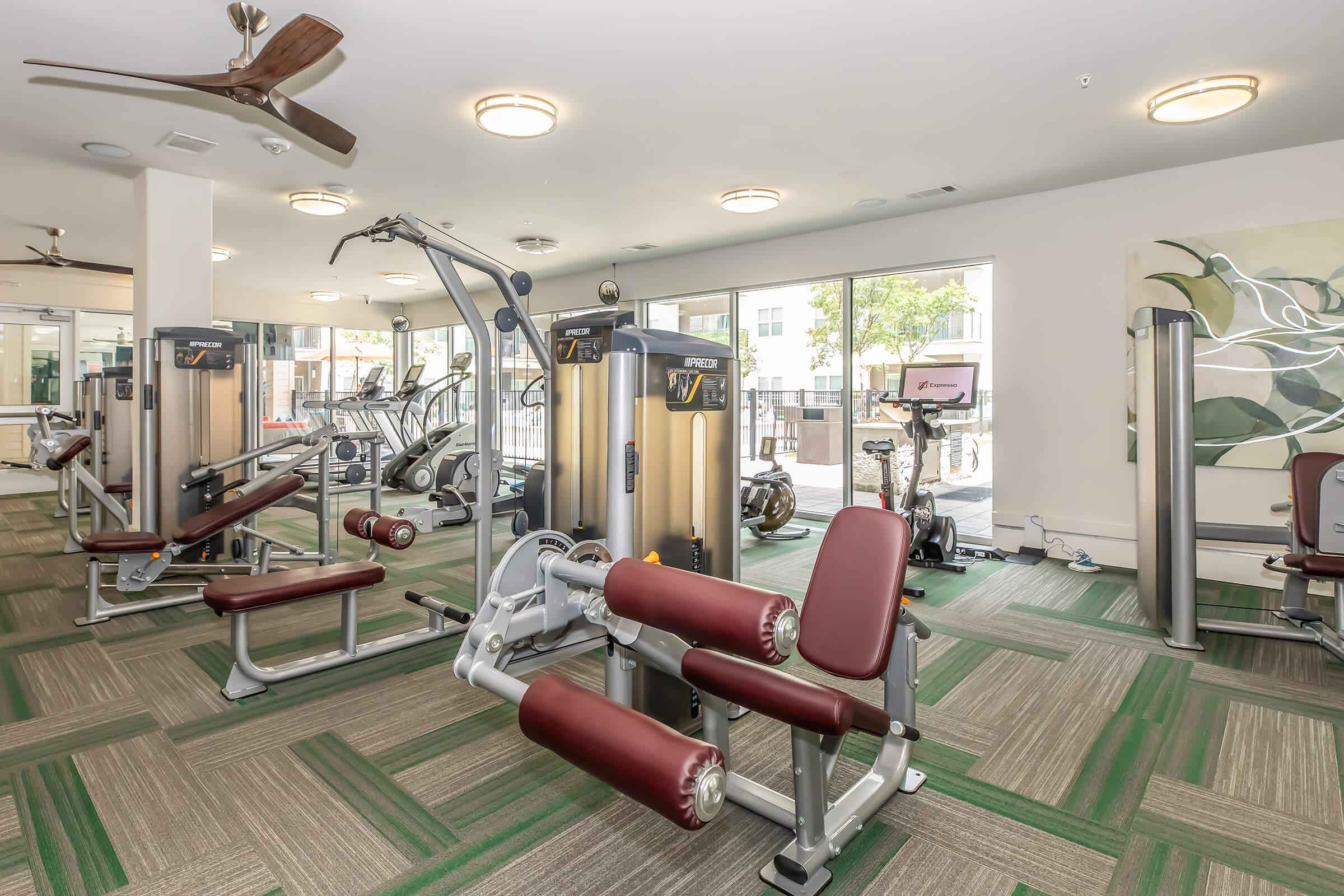 A modern gym interior featuring various exercise equipment, including weight machines, stationary bikes, and a multifunction workout machine. The space is well-lit with natural light from large windows and has a contemporary design, highlighted by green and brown flooring patterns.