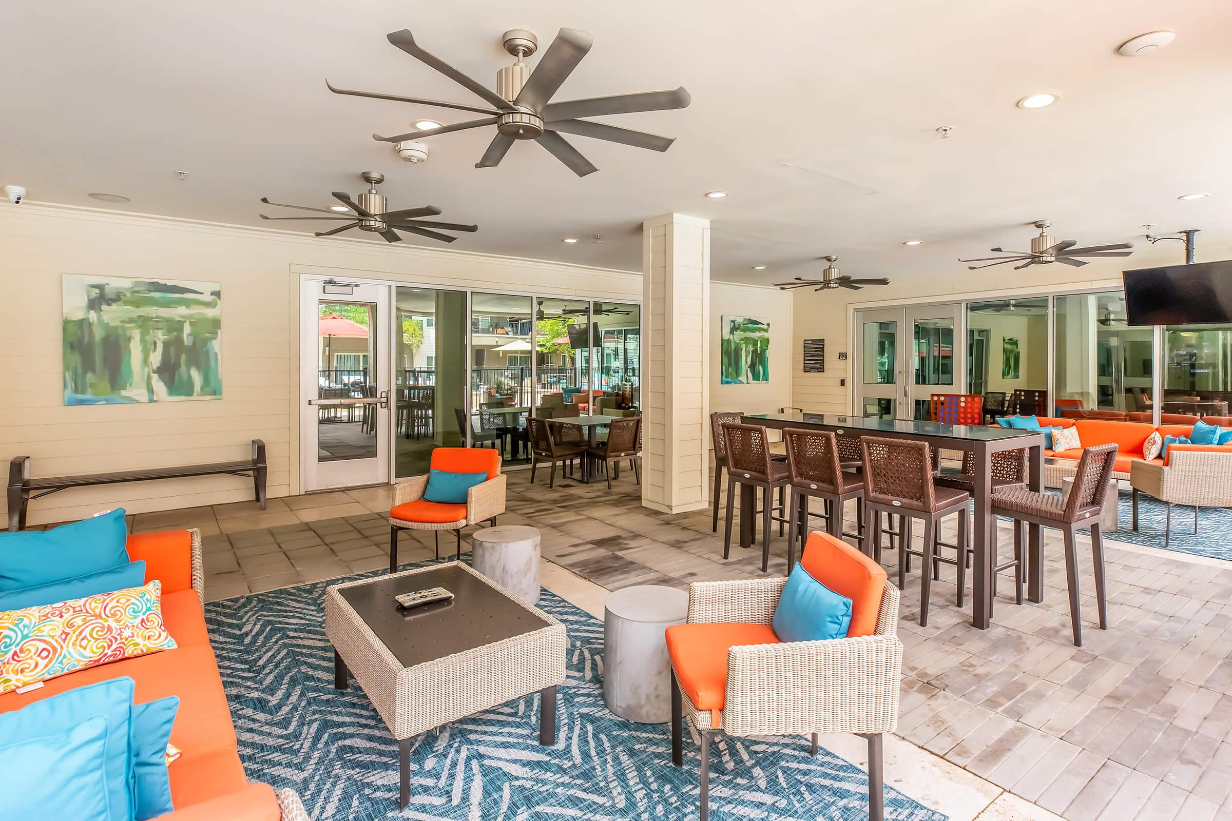 A modern outdoor lounge area featuring comfortable seating with colorful cushions, a large coffee table, and dining tables. Ceiling fans are visible, and there are large windows and light-colored walls. The space is well-lit, inviting, and designed for relaxation and social gatherings.