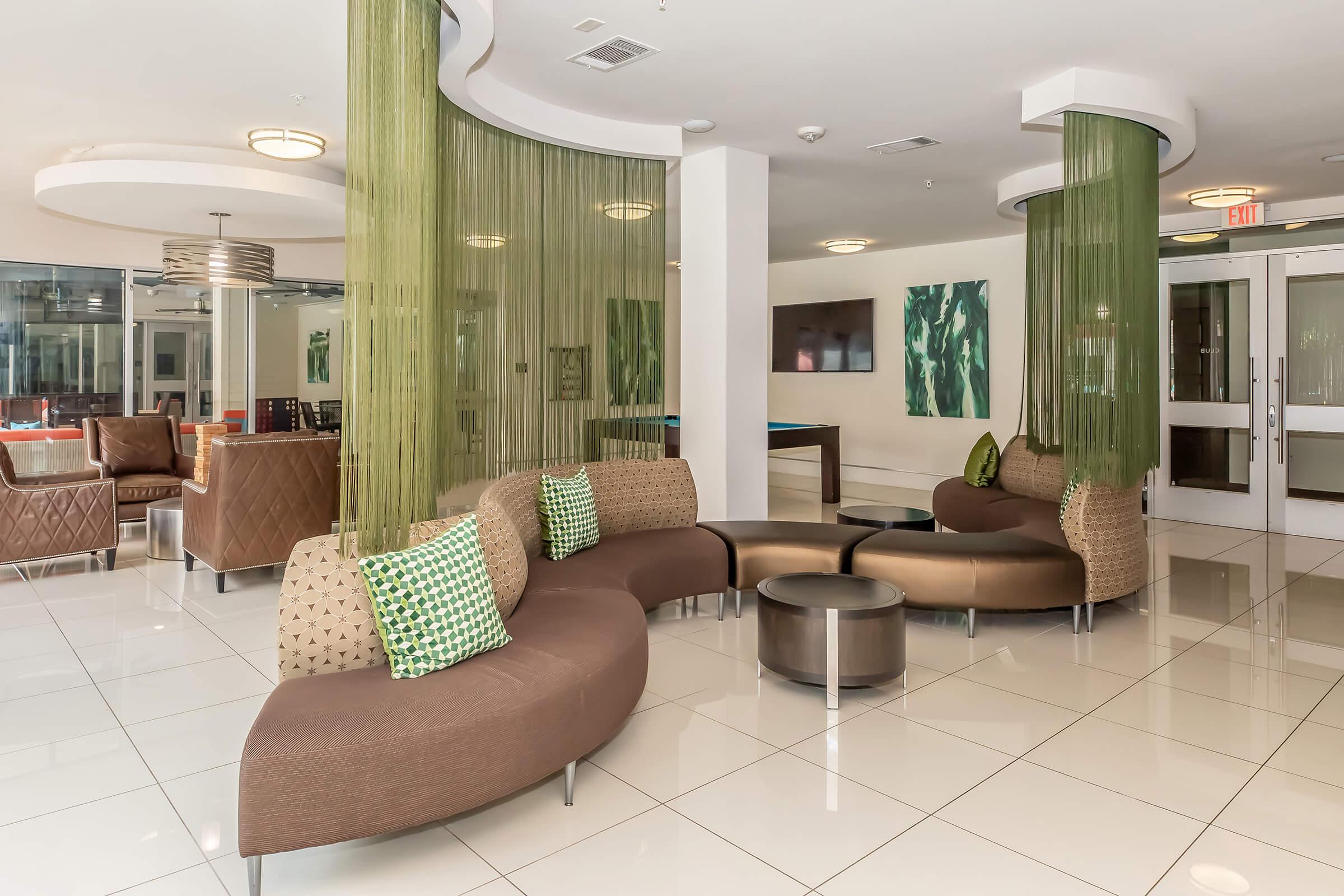 A modern lounge area featuring curved brown seating with patterned cushions, surrounded by sheer green dividers. The space includes a small round table and large windows that let in natural light, along with contemporary artwork on the walls. Stylish design elements create a welcoming atmosphere.