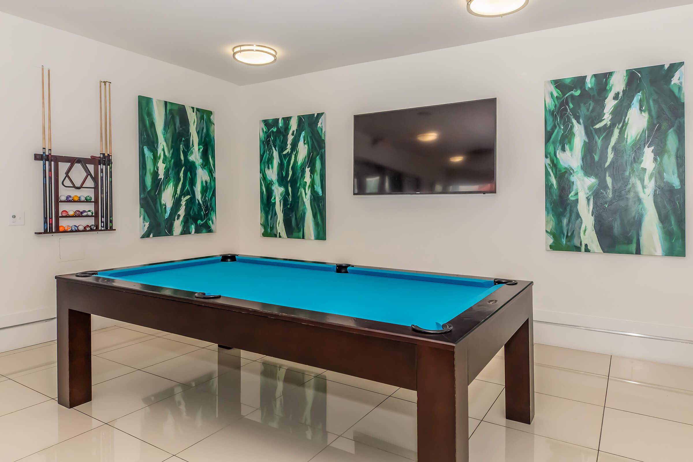 A modern game room featuring a blue felt pool table at the center. The walls are adorned with vibrant green abstract art pieces, and a large flat-screen TV is mounted on one wall. The room has a sleek, contemporary design with glossy tiled flooring and bright lighting fixtures.