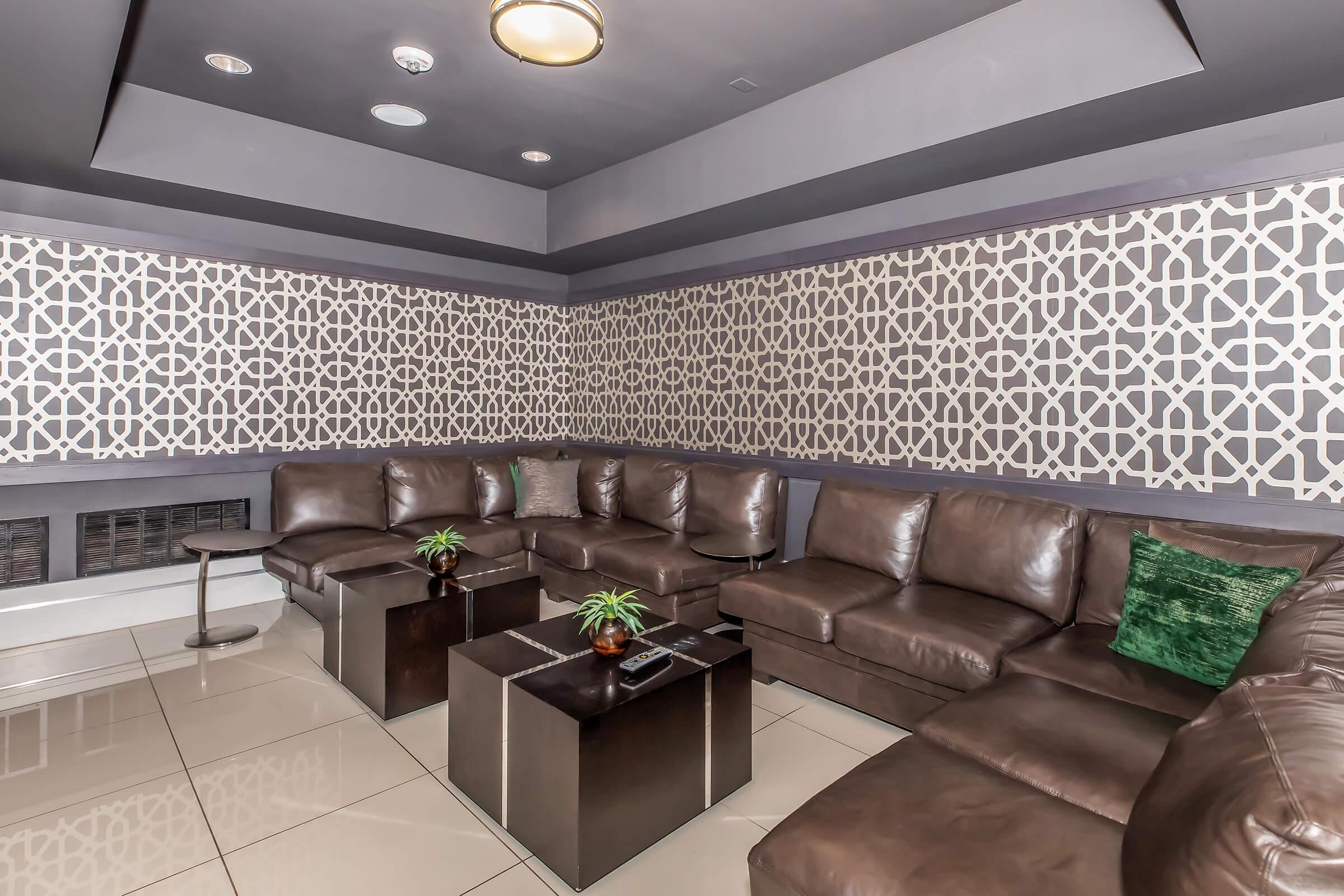 A modern lounge area featuring a brown leather sectional sofa, two glass-topped coffee tables, and decorative plants. The walls are adorned with a geometric patterned design, and the lighting is soft, creating a cozy atmosphere suitable for relaxation.