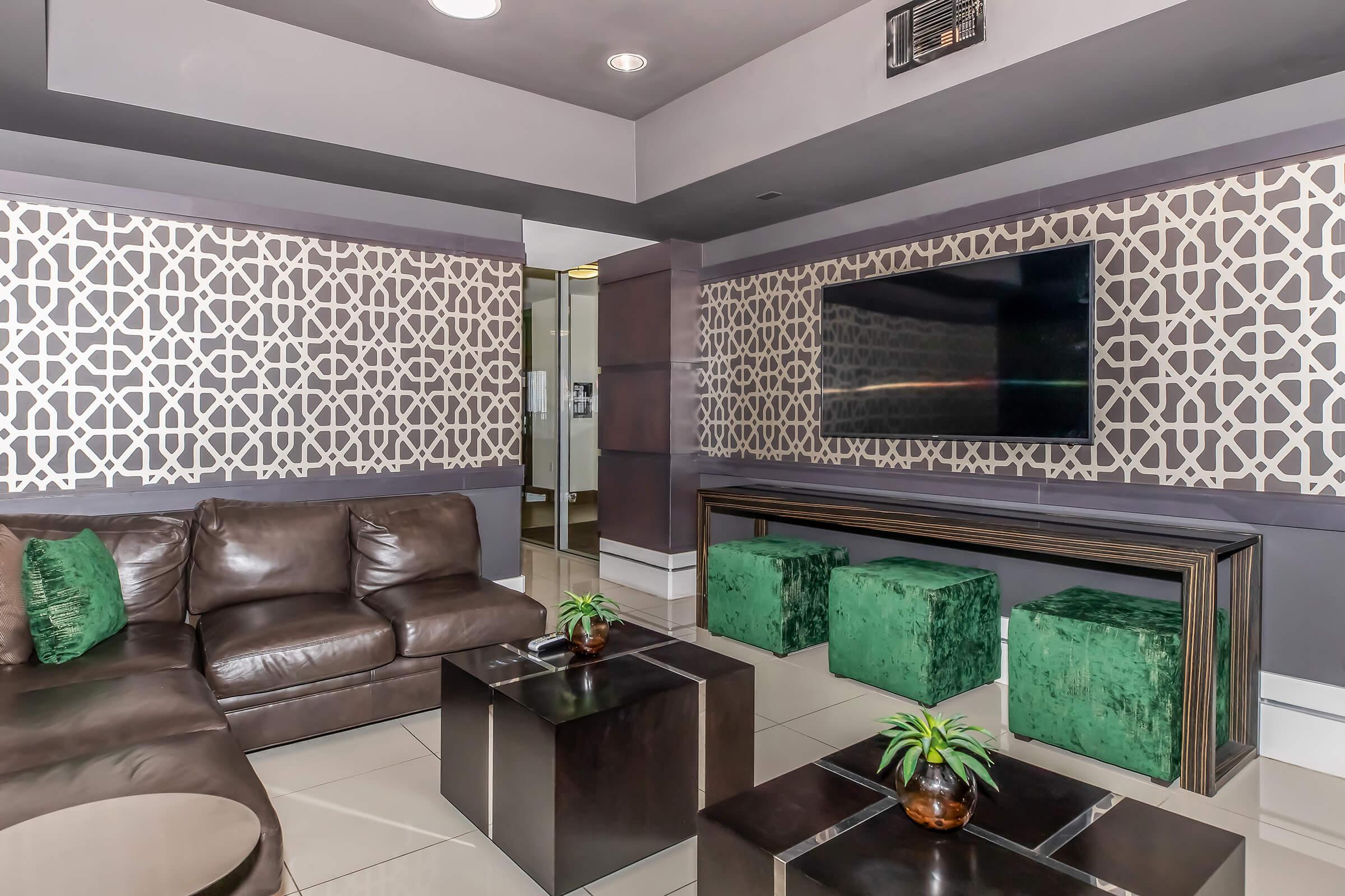 Stylish modern interior featuring a brown leather sofa, green decorative pillows, and two green velvet ottomans. A dark wood console table sits below a flat-screen TV mounted on a patterned wall. The space has sleek tiles and ambient lighting, creating a comfortable and contemporary atmosphere.