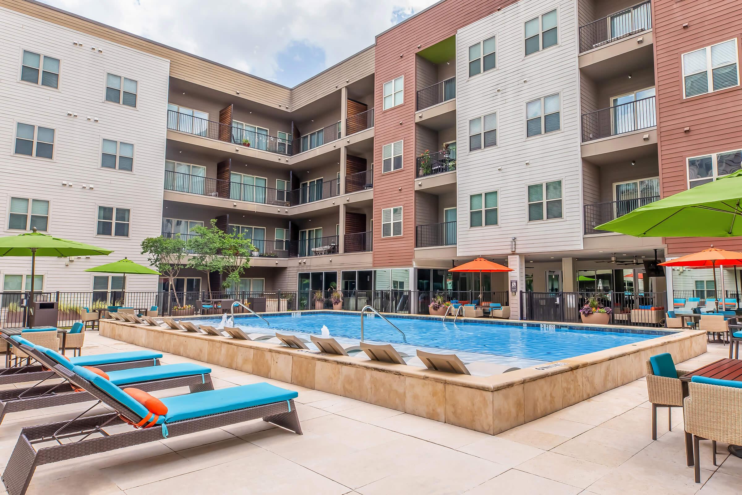A sunny outdoor swimming pool surrounded by lounge chairs and umbrellas, located in the courtyard of a modern multi-story apartment complex. The pool area features vibrant green and orange umbrellas, with residents relaxing by the water and balconies visible in the background.