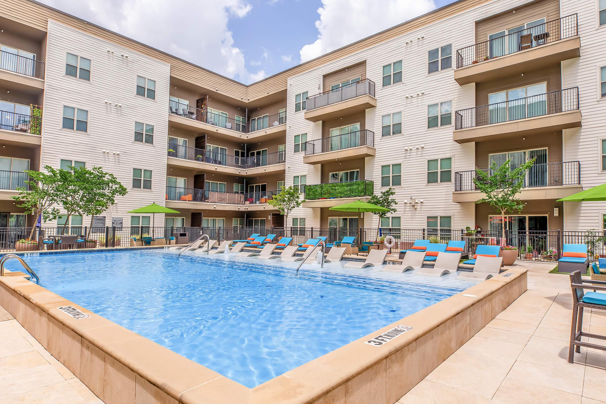 A bright, clean apartment complex featuring a swimming pool surrounded by lounge chairs and green umbrellas. The pool area is bordered by residential balconies, with lush trees and landscaping enhancing the outdoor atmosphere. A blue sky with fluffy clouds completes the scene.