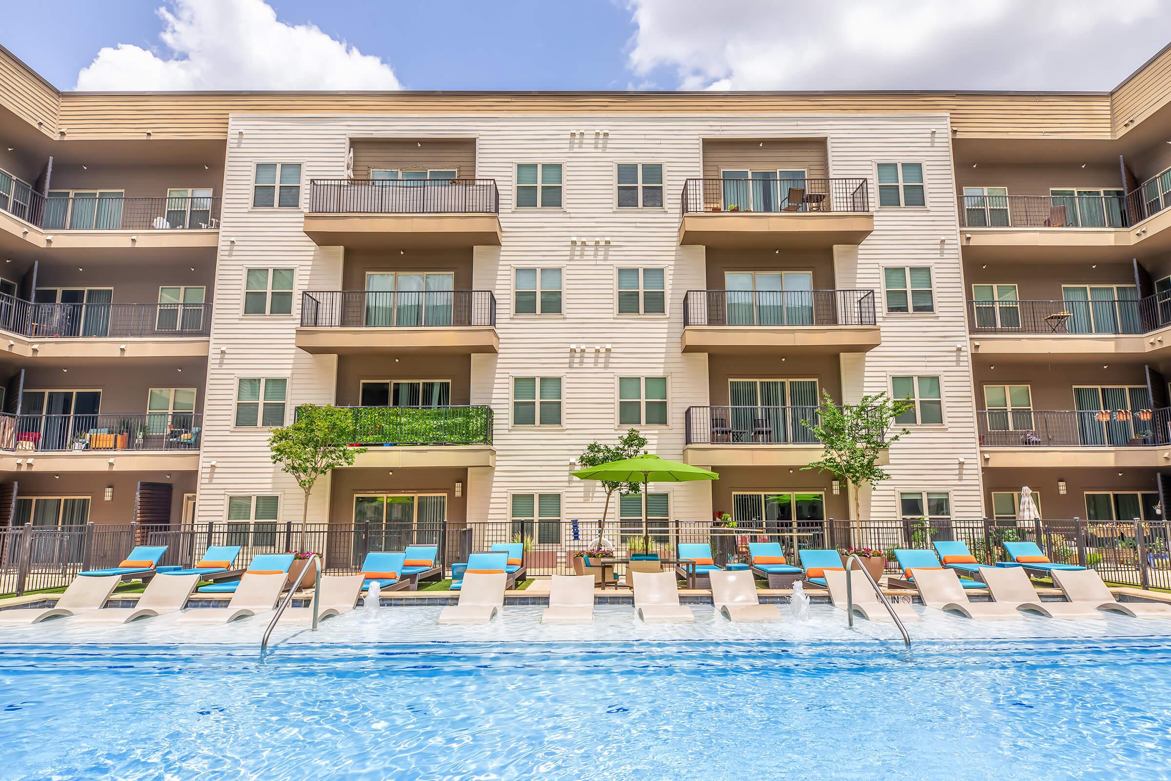 A modern apartment complex featuring multiple levels with balconies, overlooking a swimming pool. The pool area is lined with lounge chairs and umbrellas, surrounded by greenery. Bright blue skies with some clouds are visible above the building.
