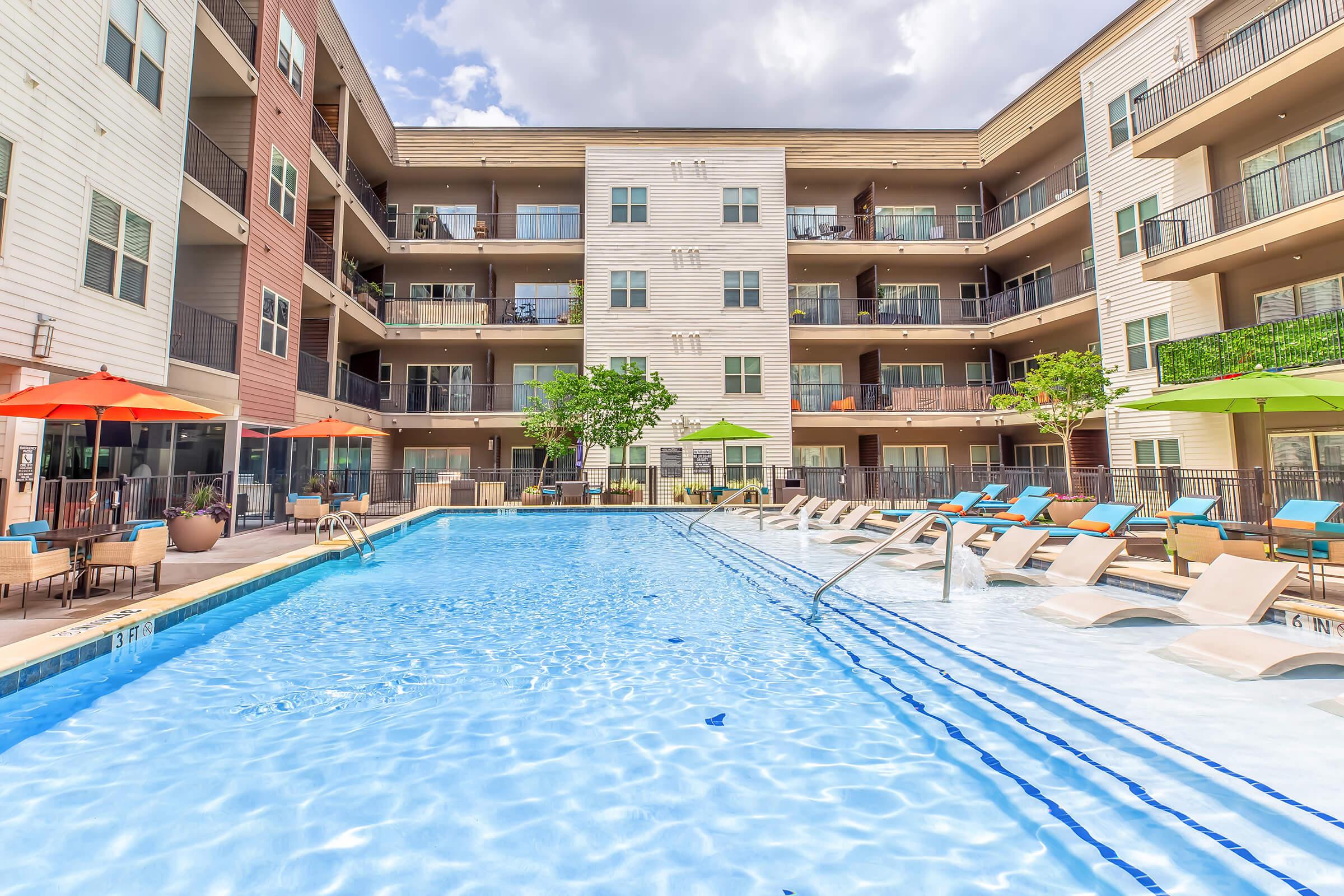 A vibrant outdoor swimming pool surrounded by modern apartment buildings. Lounge chairs line the poolside, and colorful umbrellas provide shade. Lush greenery enhances the inviting atmosphere, while sunny skies suggest a perfect day for relaxation and leisure.