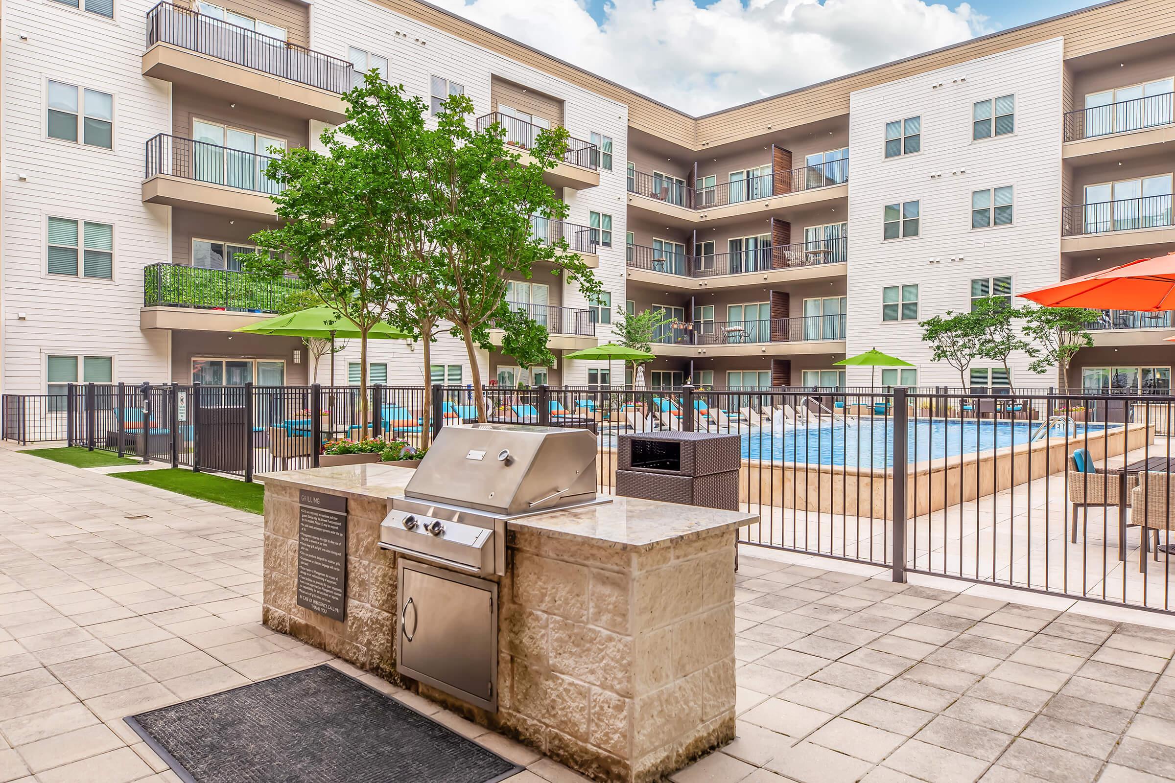 A modern outdoor area featuring a barbecue grill, surrounded by a fenced pool, lounge chairs, and green umbrellas. The backdrop includes an apartment building with multiple balconies and large windows, creating a relaxing environment for residents to enjoy.