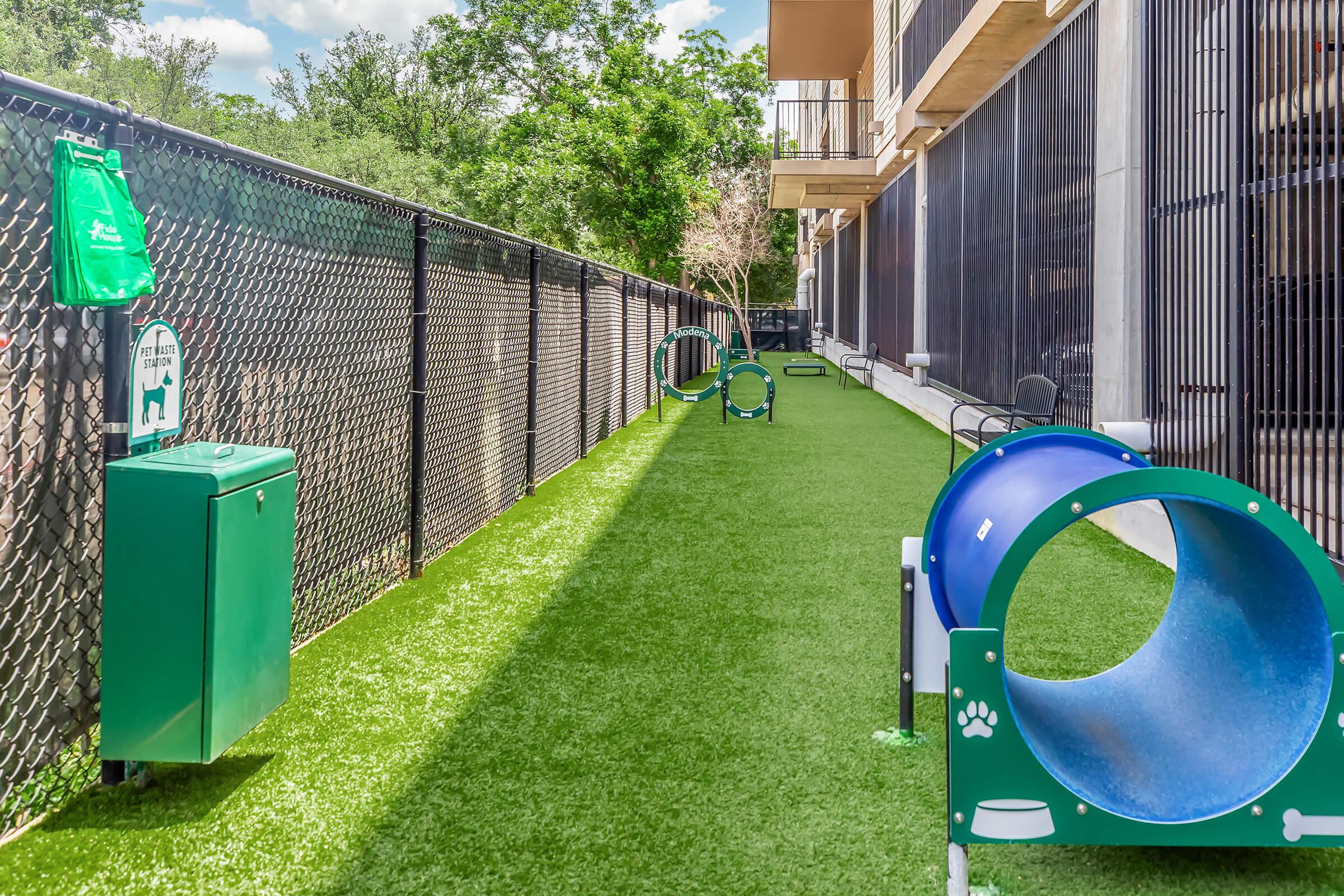 A pet-friendly area featuring a grassy pathway lined with a fence, plastic agility tunnels, and waste disposal stations. The space is designed for dogs to play and exercise, with greenery and natural light in a residential setting.