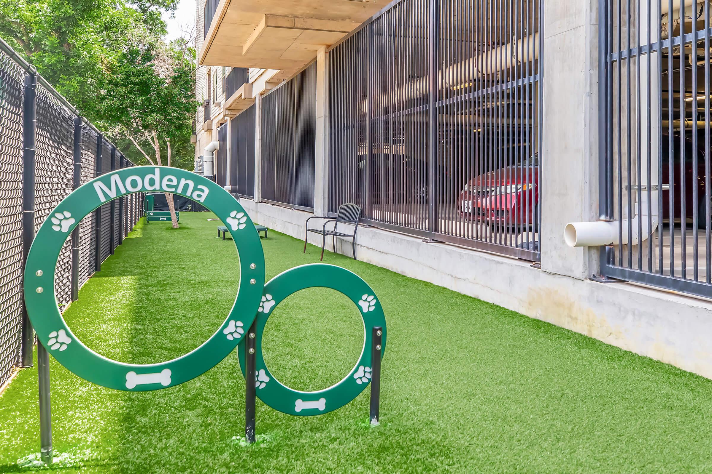 Green dog agility equipment with the name "Modena" displayed, set in a landscaped area with artificial grass. A chair is nearby, and a parking garage is visible in the background. The space appears pet-friendly and well-maintained.