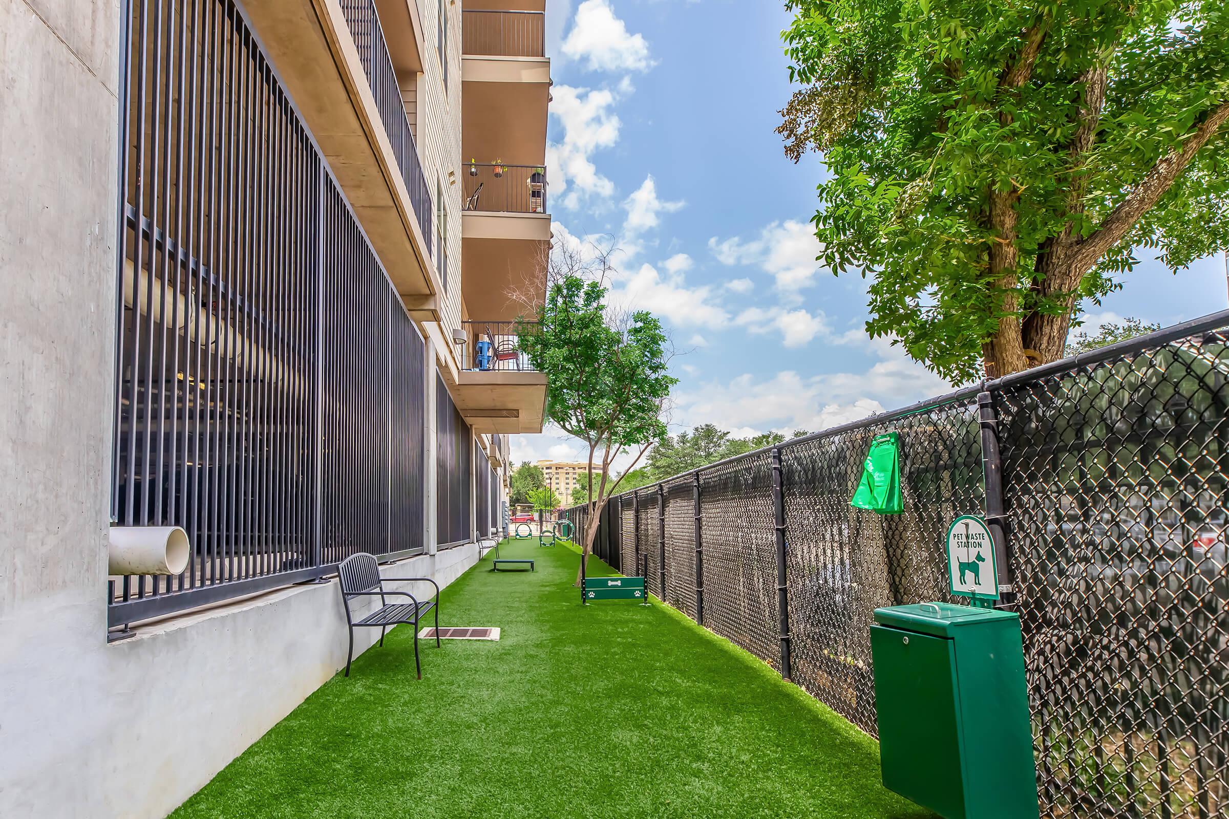 A green, artificial turf area adjacent to a modern building. The space features benches, a waste disposal bin, and a small fenced section with trees. It is set against a backdrop of blue skies and scattered clouds, providing a pleasant outdoor environment.