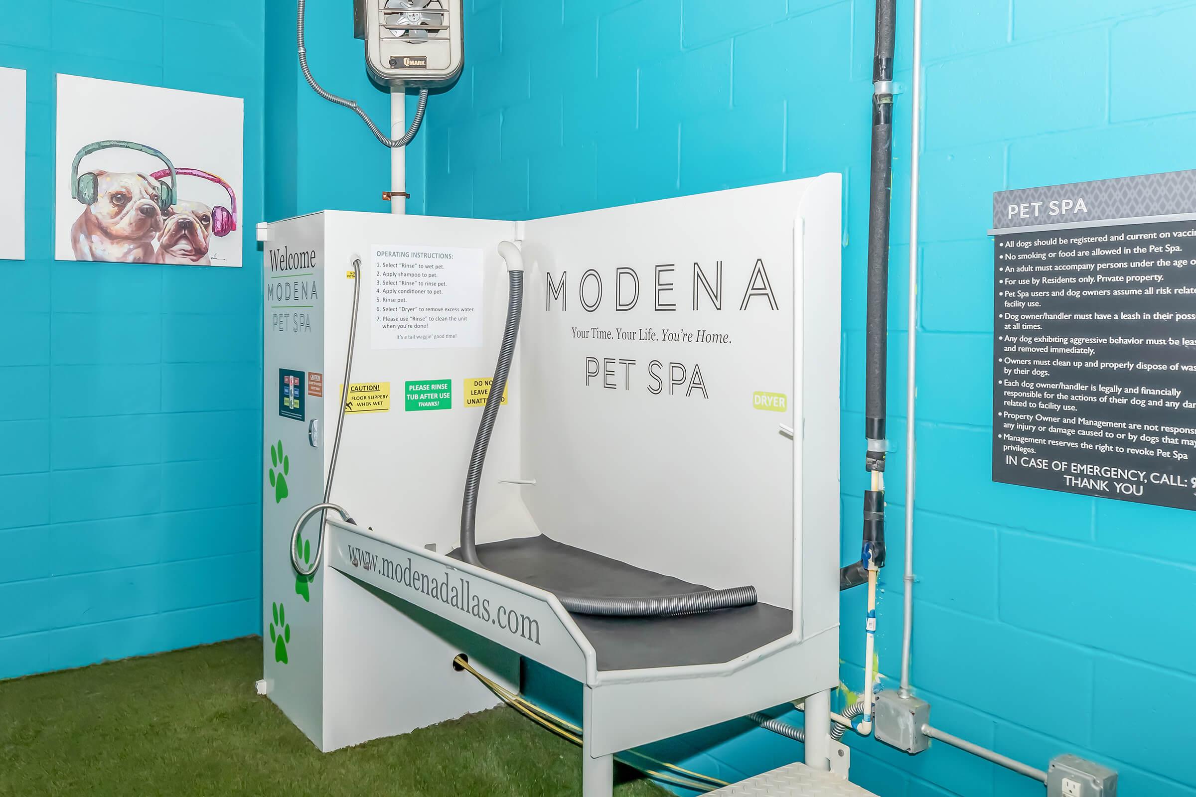 A clean and modern pet spa facility featuring a bathing station with a non-slip surface, surrounded by a bright blue wall. The area includes a sign with instructions and a website link for more information, as well as emergency contact information. The floor is covered with green grass-like material for a playful atmosphere.