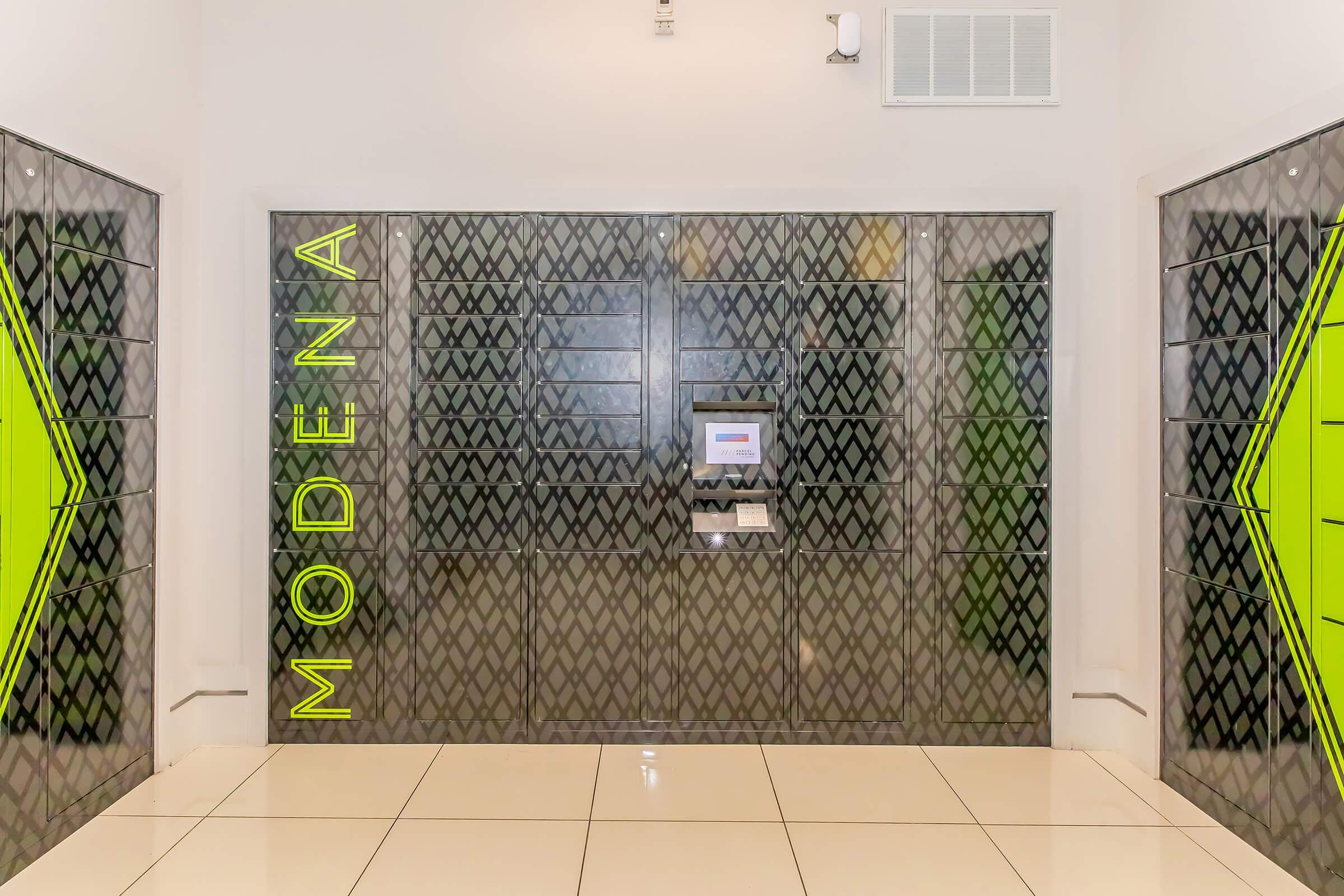 A modern, sleek storage or locker area with a glass façade, featuring a geometric pattern. The word "MODENA" is displayed prominently in bright green on the left side. A digital interface or control panel is visible at the center, set against the reflective surface. The floor is light-colored, complementing the contemporary design.