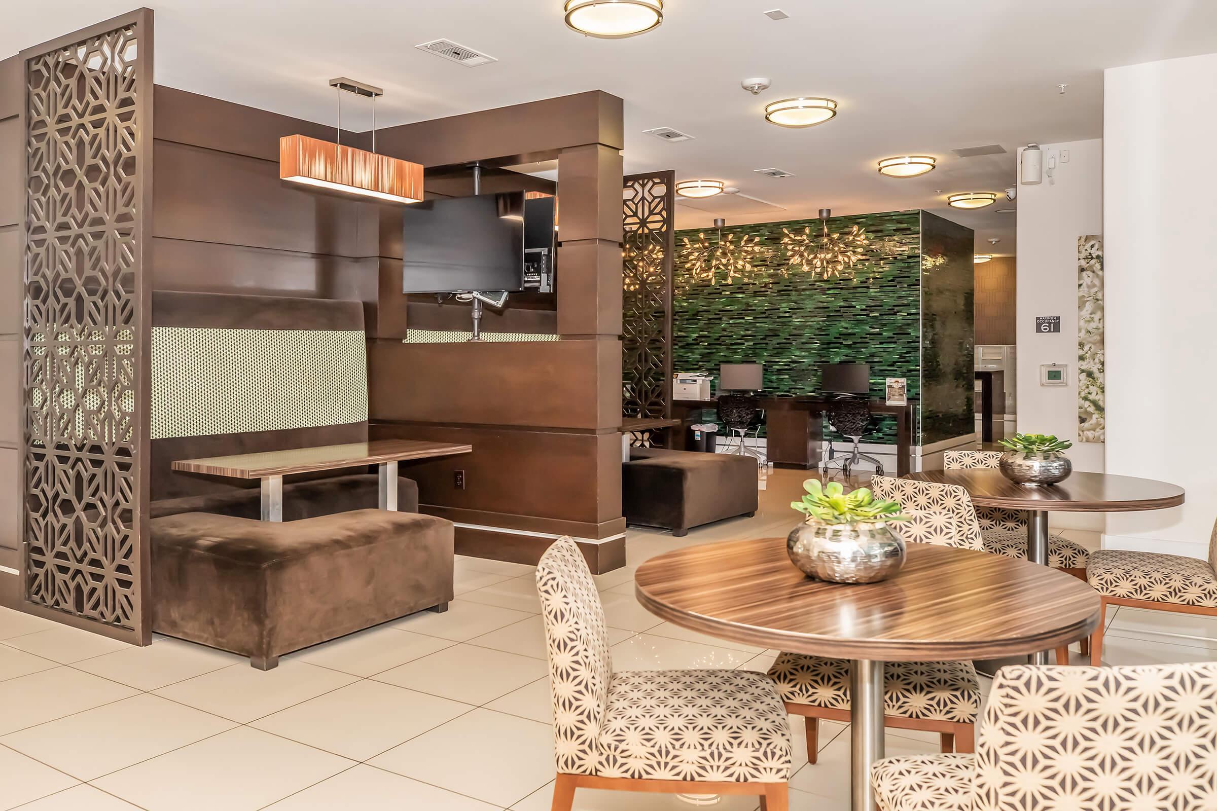 Interior view of a modern lounge area featuring decorative partitions, wooden furniture, and green accent walls. Round tables with stylish chairs are arranged around the space, complemented by potted plants. A flat-screen TV is mounted on the wall, enhancing the contemporary design.