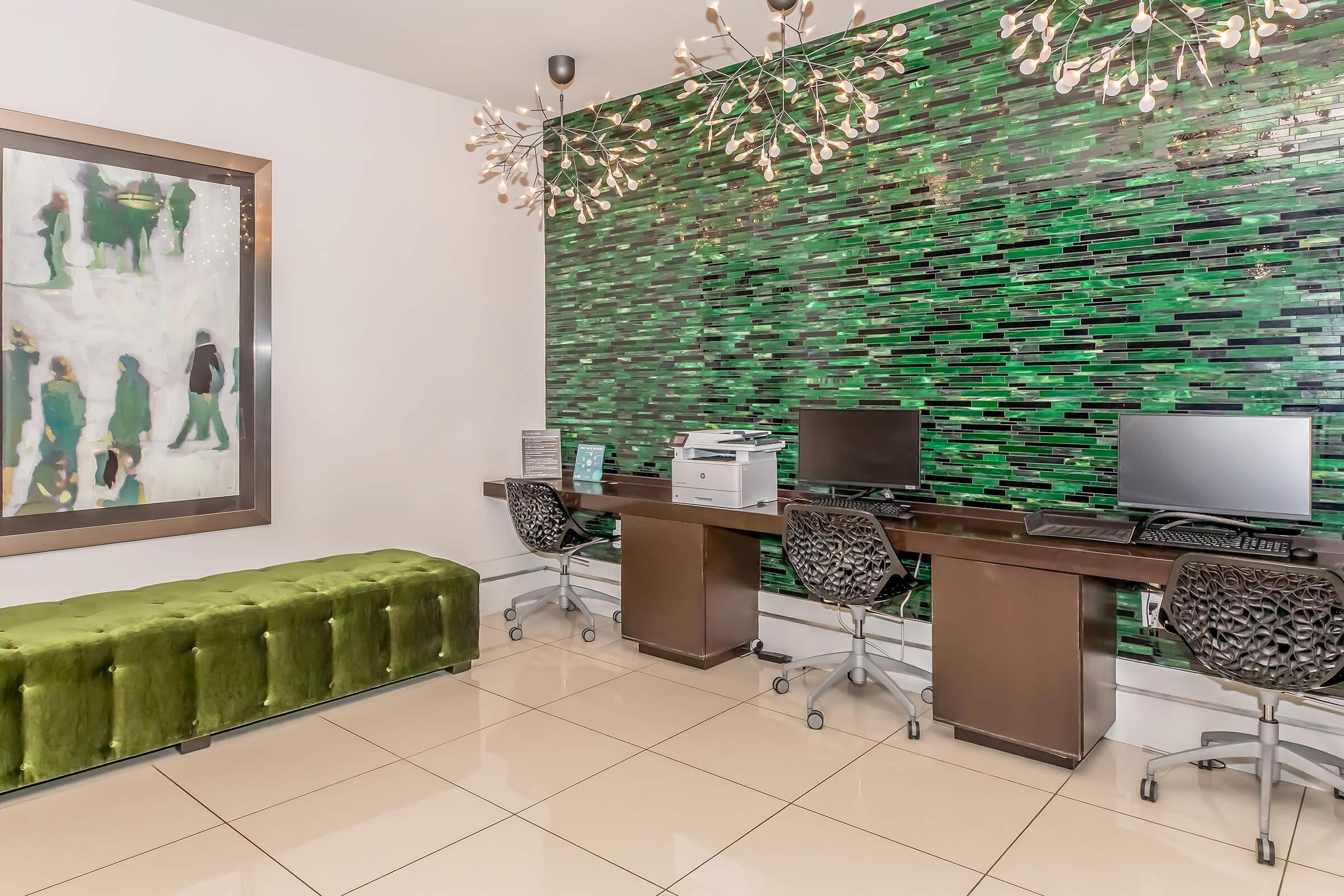 A modern workspace featuring two desktop computers with black chairs, a printer, and a green velvet bench. The backdrop consists of a vibrant green mosaic wall, complemented by artistic light fixtures hanging from the ceiling, creating a stylish and contemporary atmosphere.