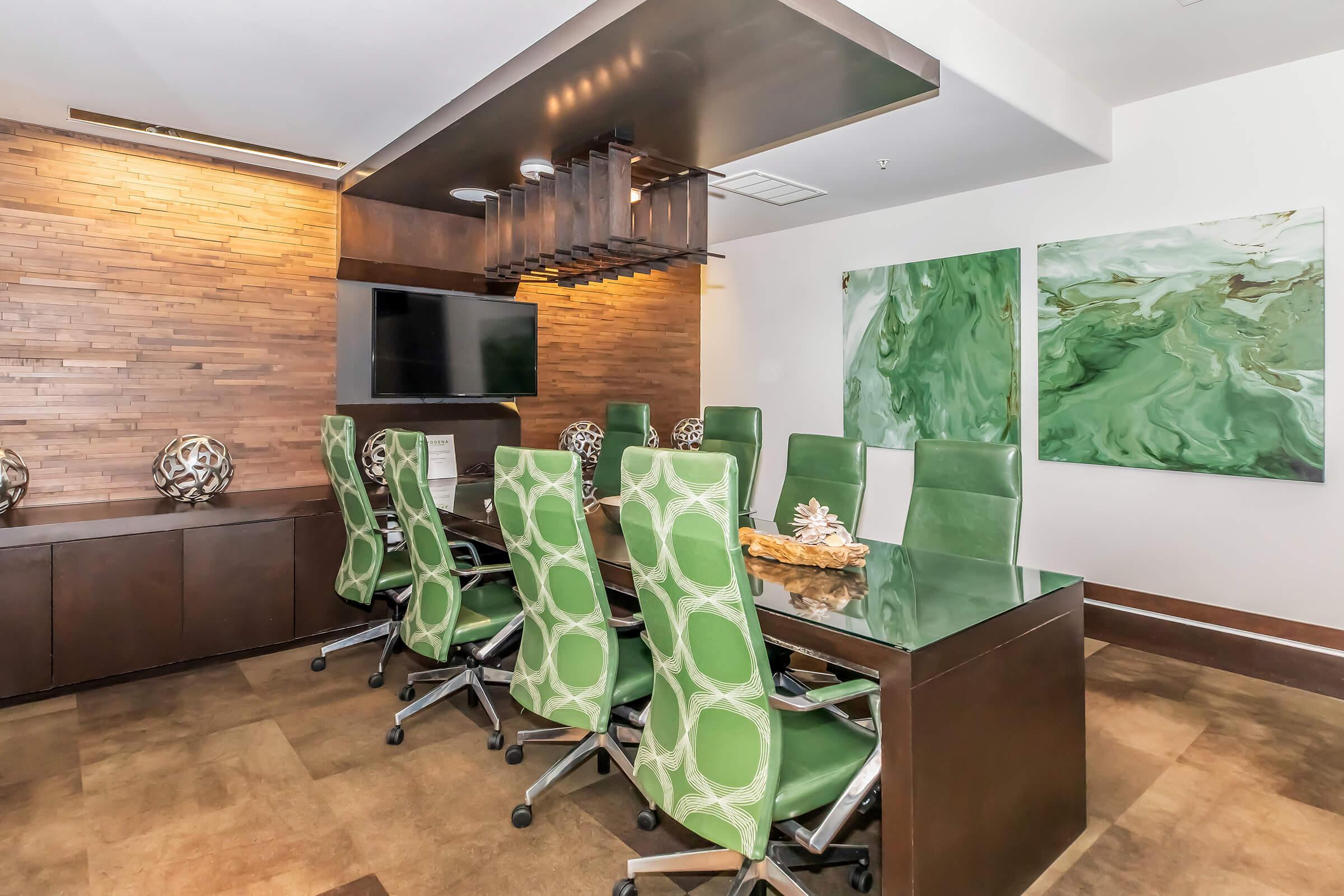 A modern conference room featuring a large glass table surrounded by green and white patterned chairs. The room is decorated with wooden paneling and includes abstract green artwork on the walls. There is a flat-screen TV mounted and distinct lighting fixtures above the table, creating a professional environment.
