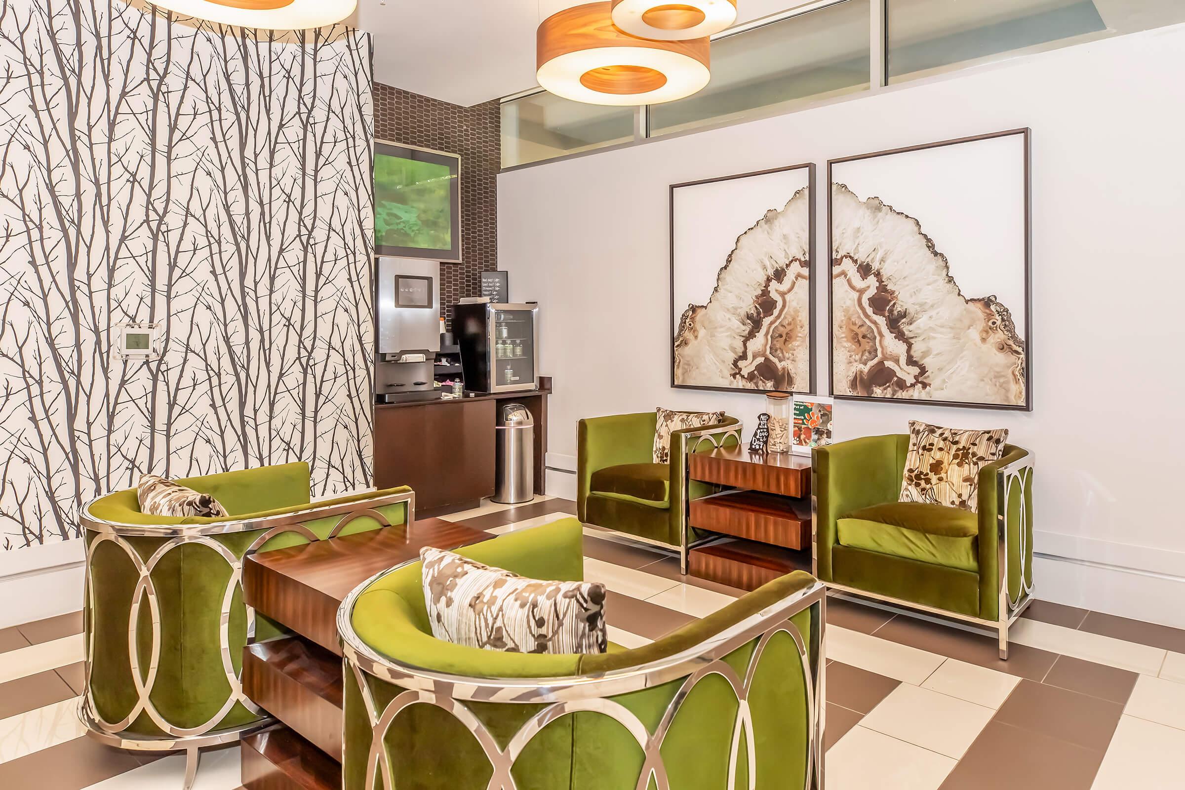 A modern lounge area featuring two green armchairs with decorative pillows, a wooden coffee table, and two large framed artworks. The wall has a nature-inspired design with tree motifs, and there is a beverage station in the background. The flooring alternates between light and dark tiles.