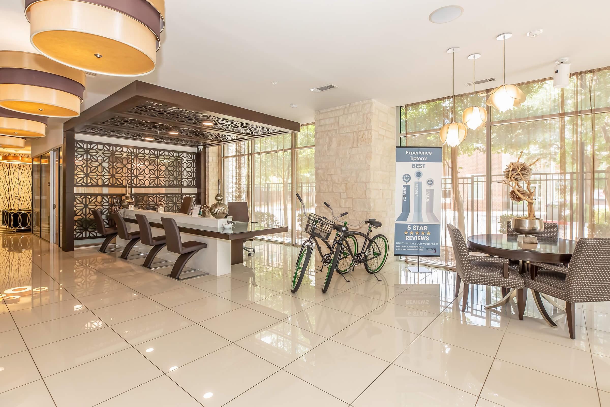 Modern lobby with stylish decor featuring a large stone wall, elegant lighting fixtures, and a dining area with dark chairs around a long table. Two bicycles are parked beside the wall, and there is a banner displaying five-star reviews. Floor tiles are glossy and reflect the interior design.
