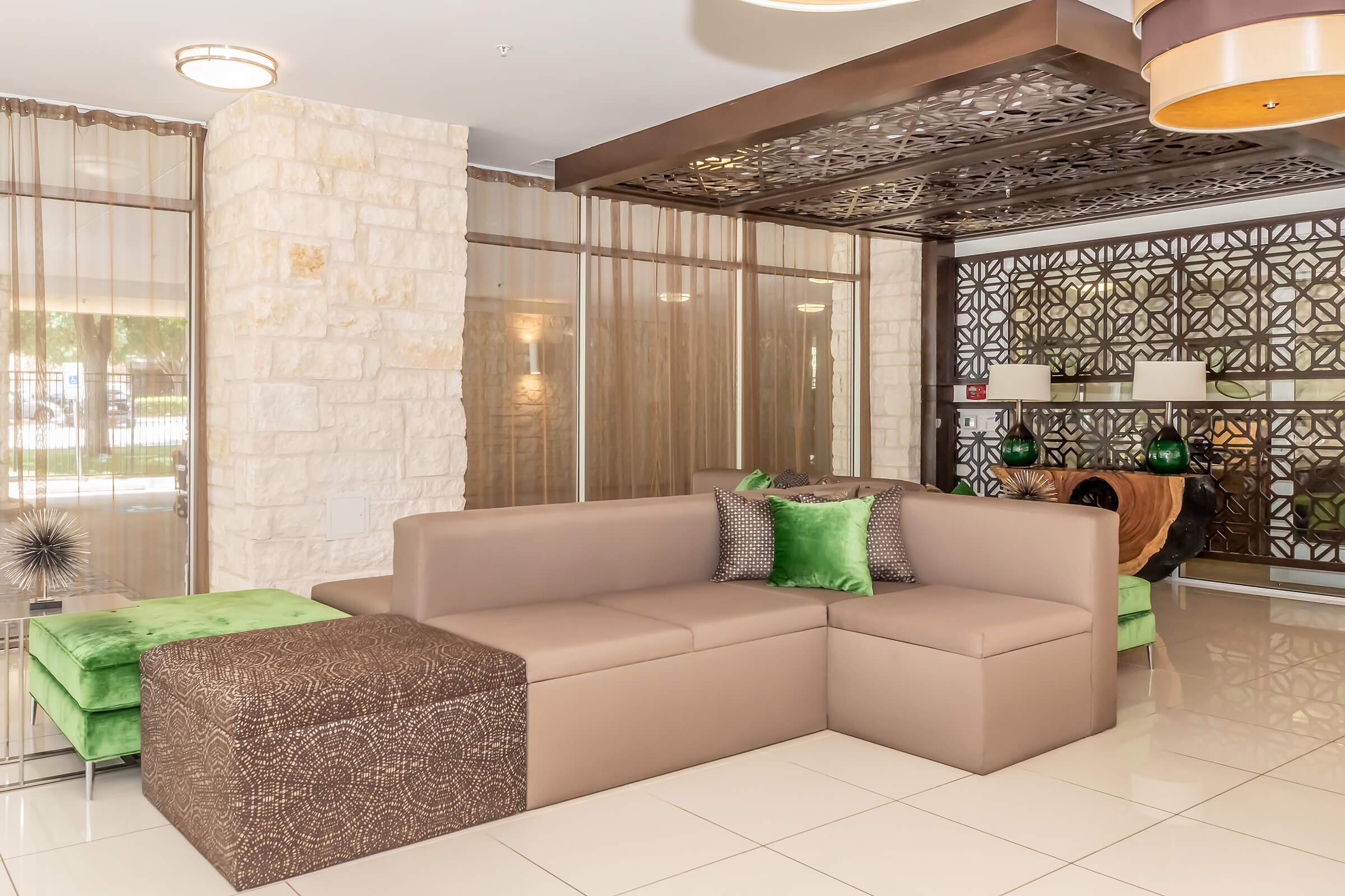 Modern lounge area featuring a sectional sofa in light brown and a brown patterned ottoman. The space is accented with green cushions and decorative plants. Elegant lighting and textured wall designs create a stylish and welcoming atmosphere, framed by sheer curtains and stone walls.