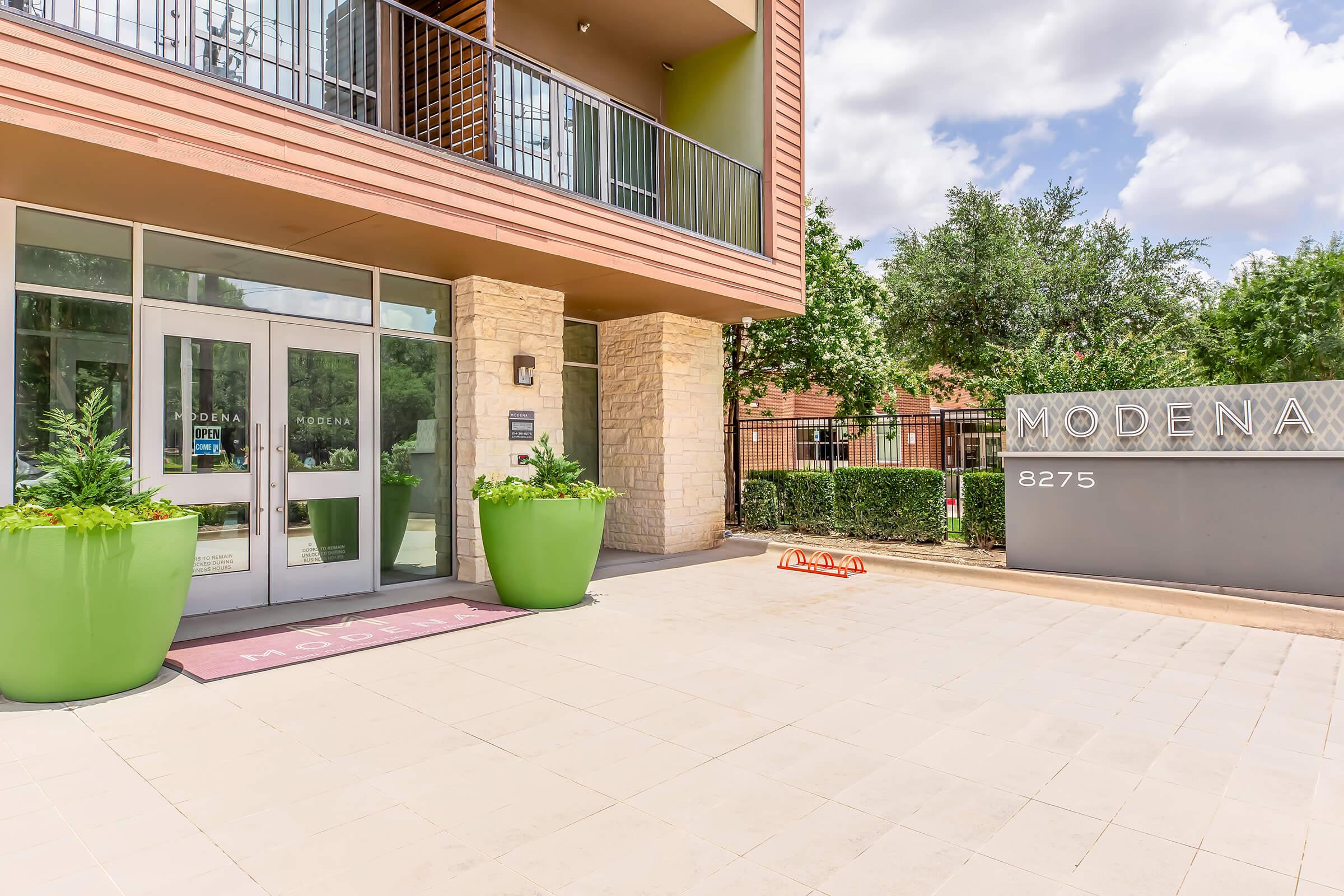 Modern apartment building entrance with large green planters, a welcome mat, and a sign reading "Modena" alongside the address "8275." The area is well-lit and features trees in the background, creating a welcoming atmosphere.